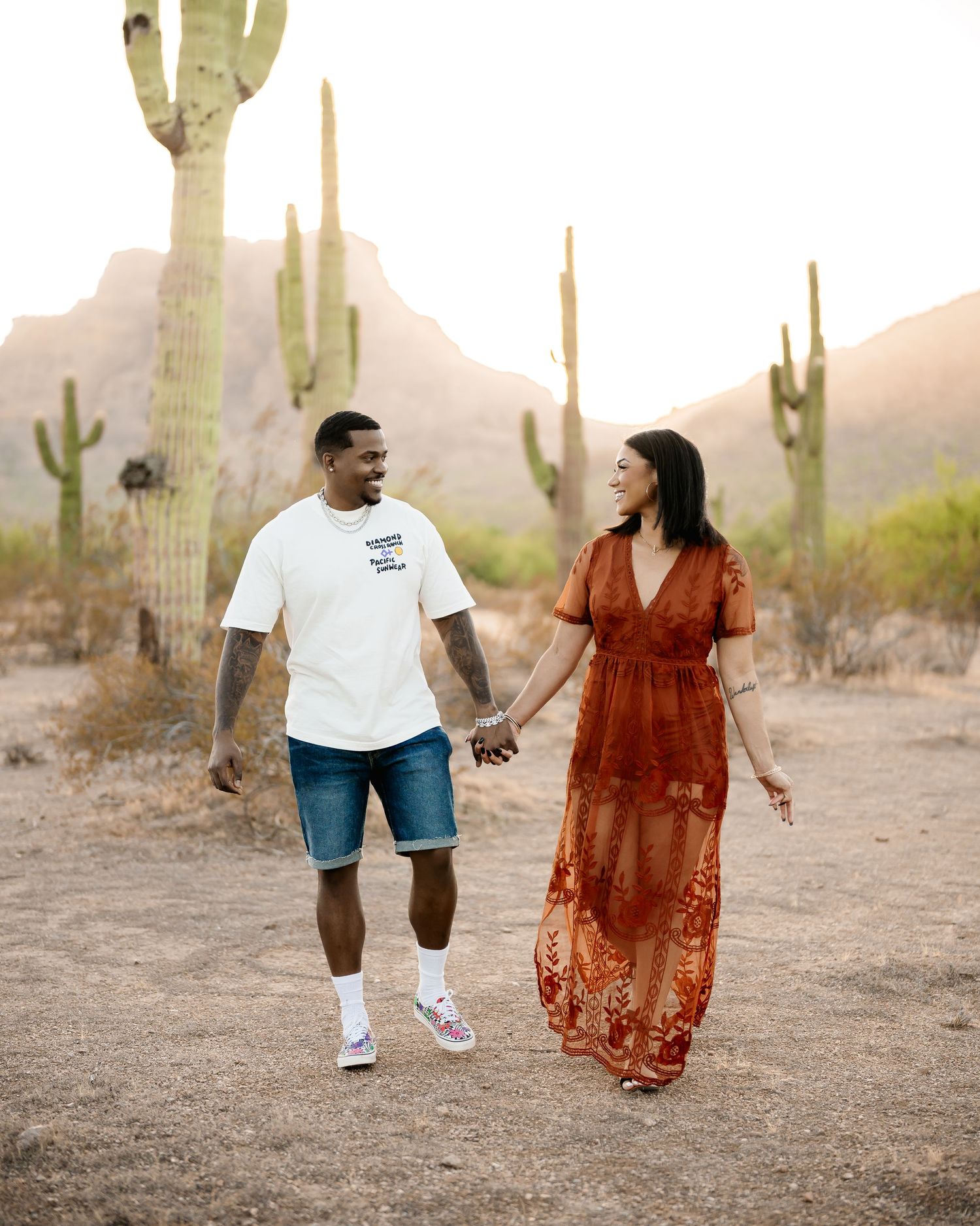 Couple walking hand in hand through desert landscape with towering saguaro cacti.