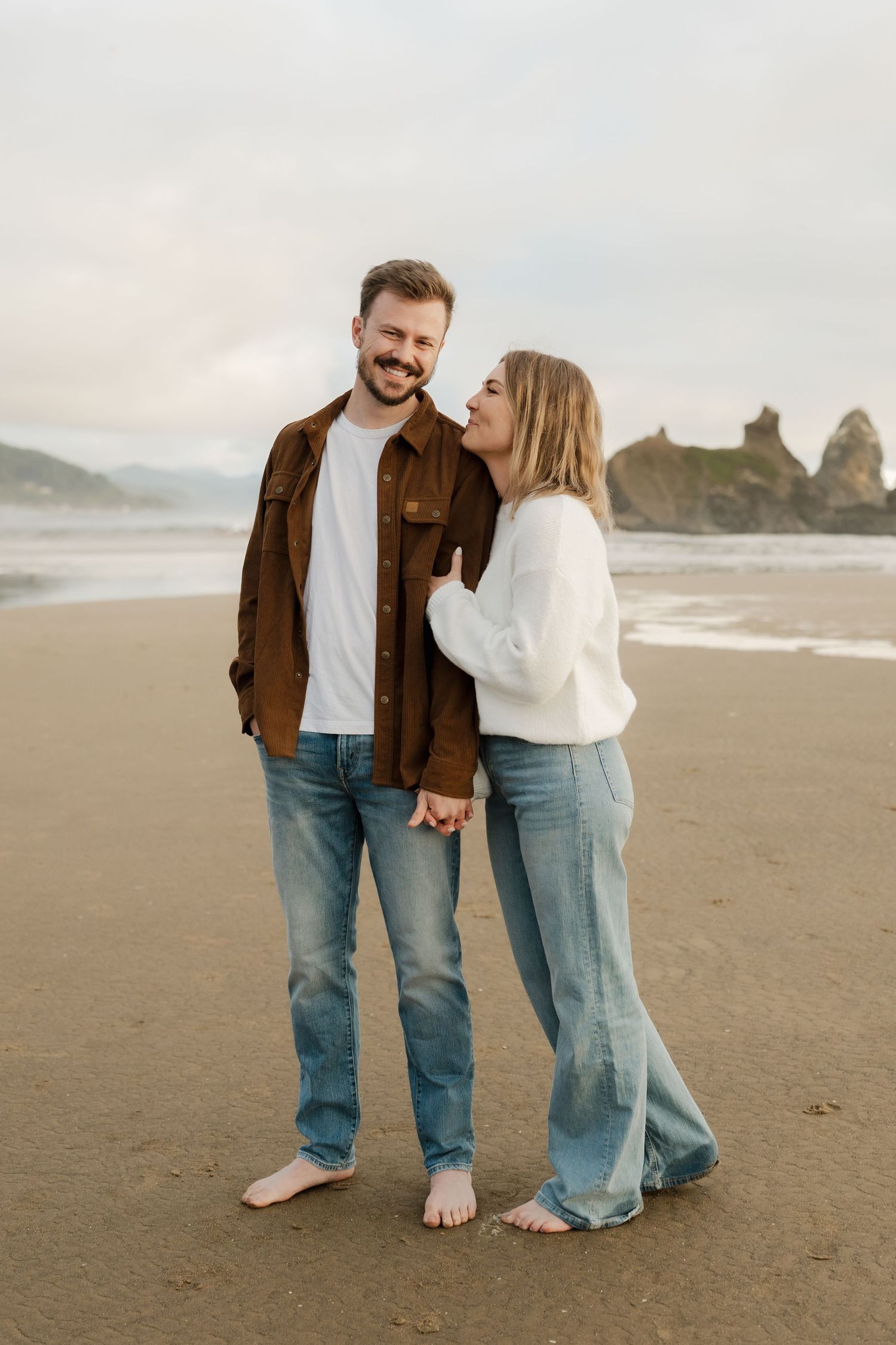 A couple holds hands and smiles on a sandy beach, dressed casually in jeans and sweaters with ocean rocks behind.
