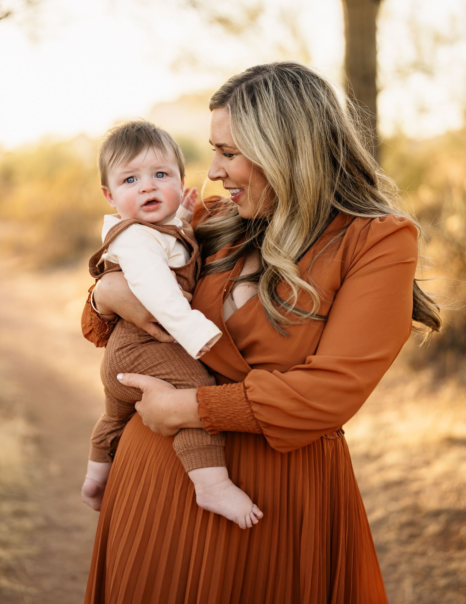 Mother in rust-colored dress holding infant in desert setting.