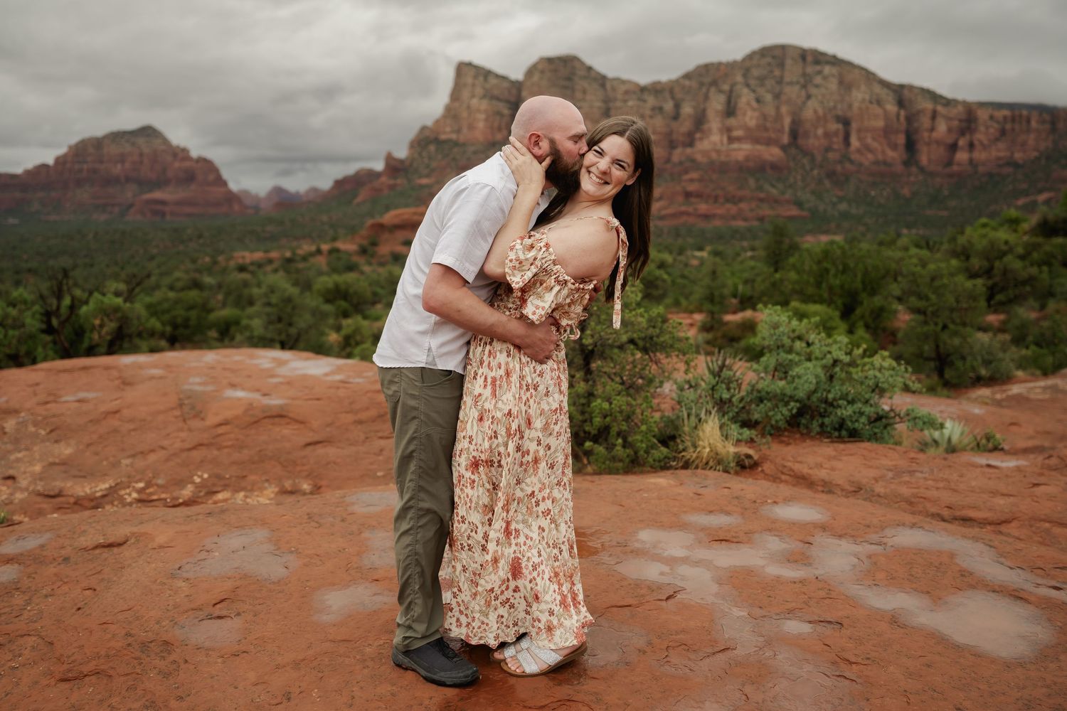 A couple shares intimate moments on red rocks with towering desert cliffs behind them.