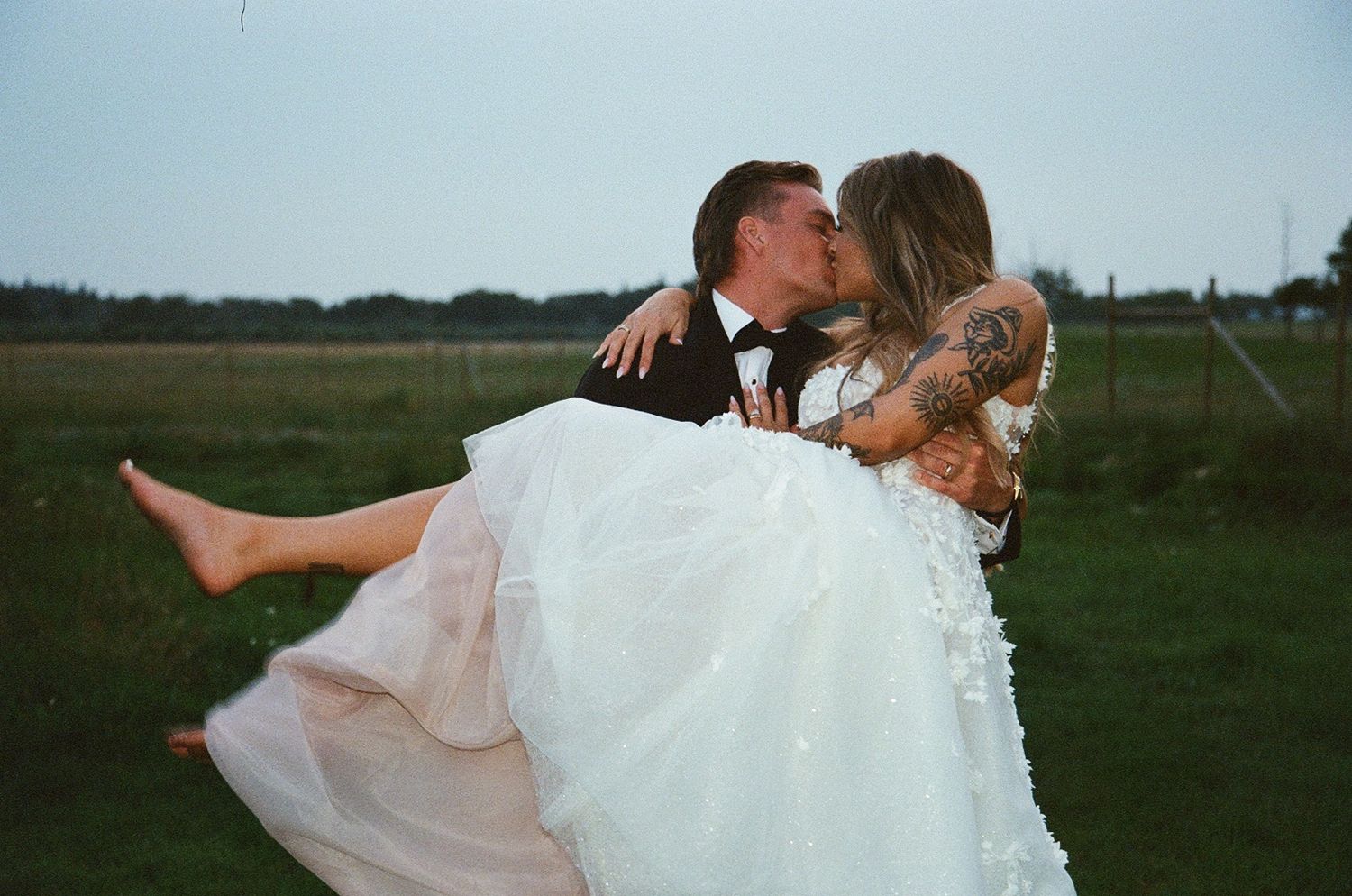 Groom carries bride in white wedding dress while sharing a kiss outdoors on lush green field at dusk.