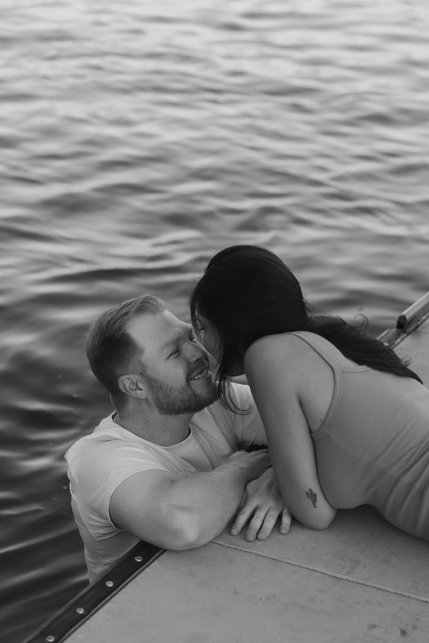 Black and white photo of a couple sharing a romantic kiss on a dock by the water.