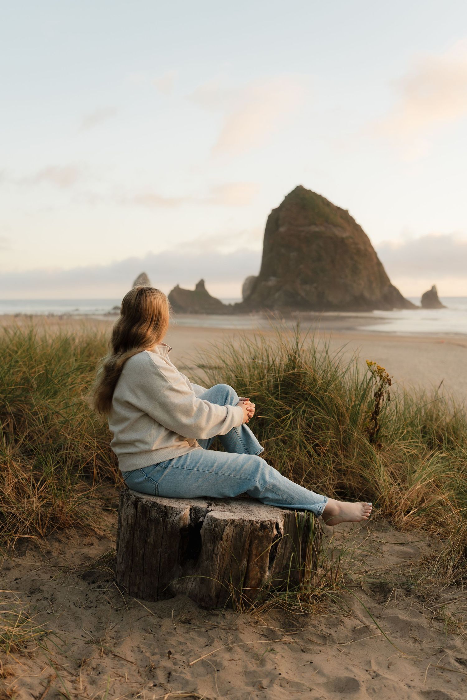 Woman sitting on a driftwood stump at Cannon Beach, Oregon, gazing at Haystack Rock during a peaceful sunset.