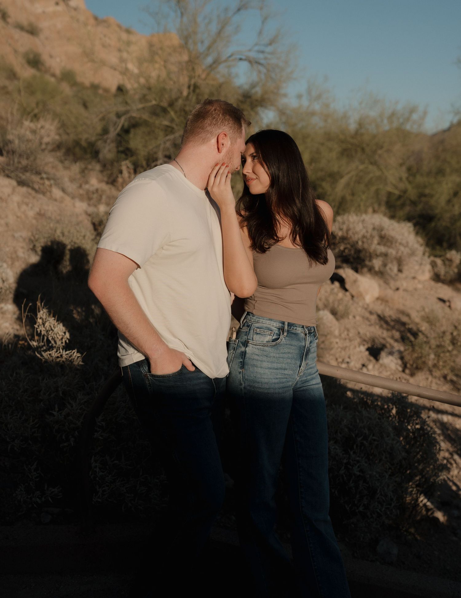 Couple sharing a romantic moment in a desert landscape with rocky hills and sparse vegetation in warm golden light.