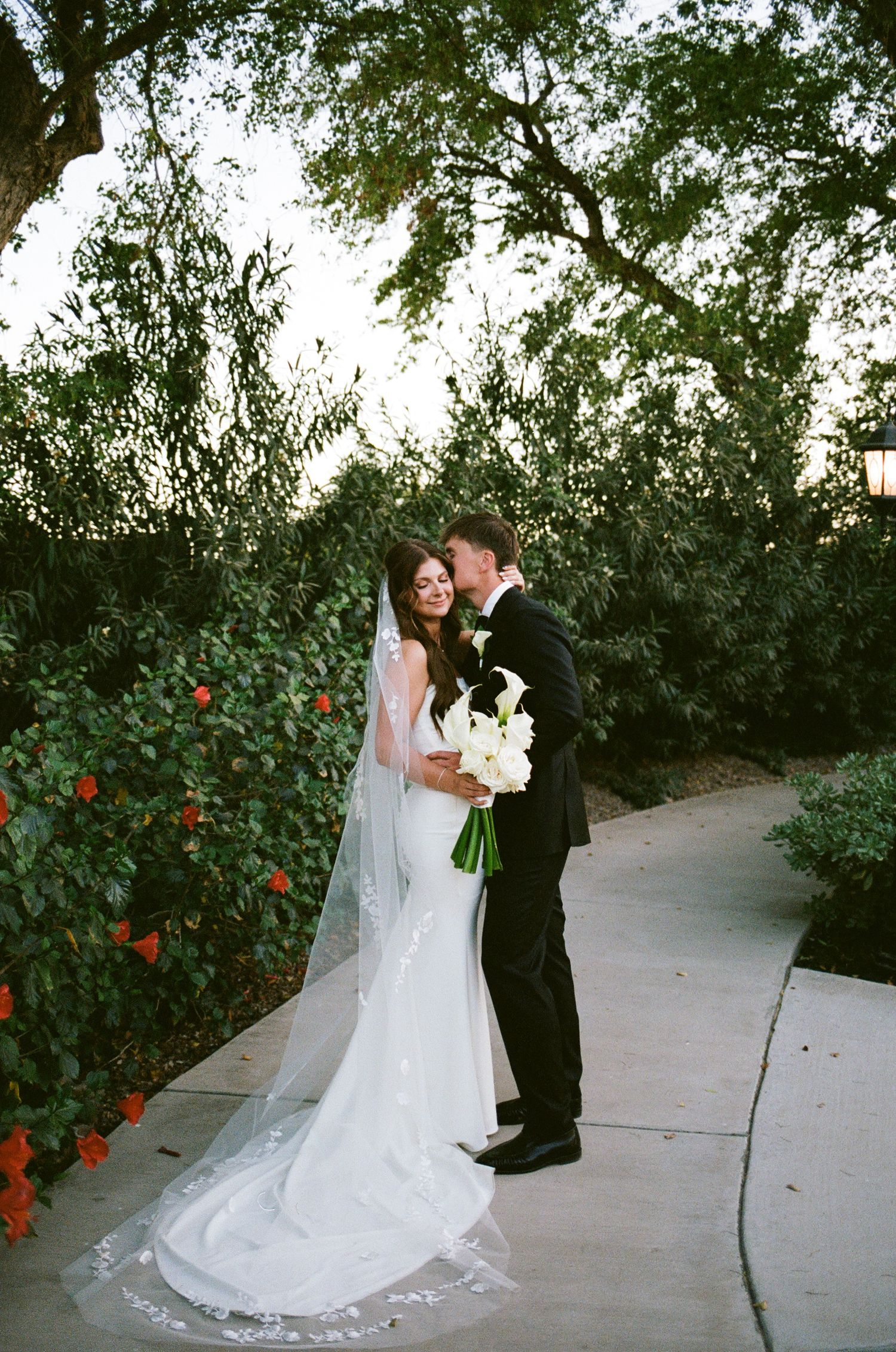 Bride in white gown with long veil holding white bouquet shares romantic moment with groom in black tuxedo on garden path.