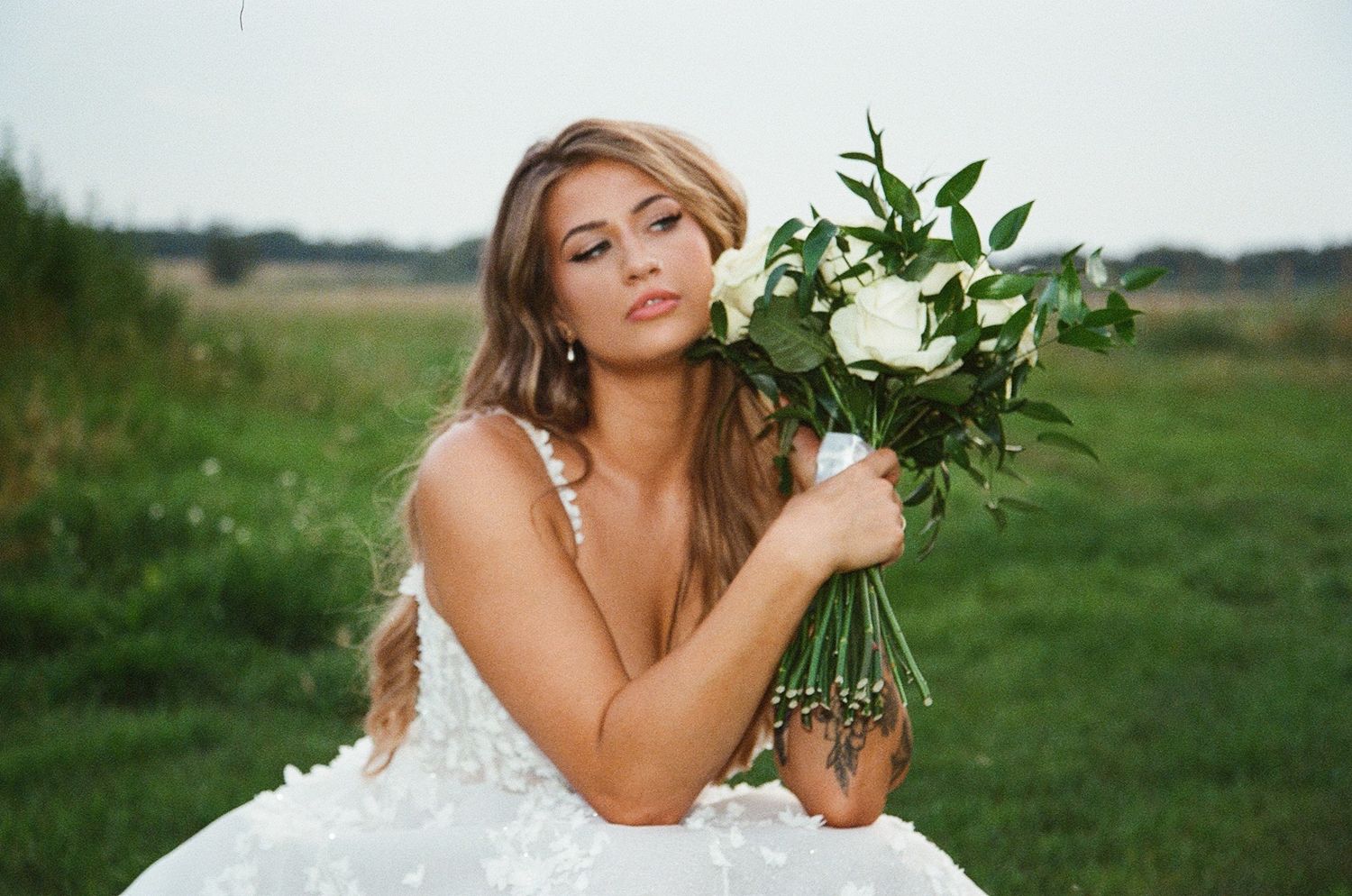 Bride in white gown holding bouquet of white roses in green field, elegant wedding photography outdoor.