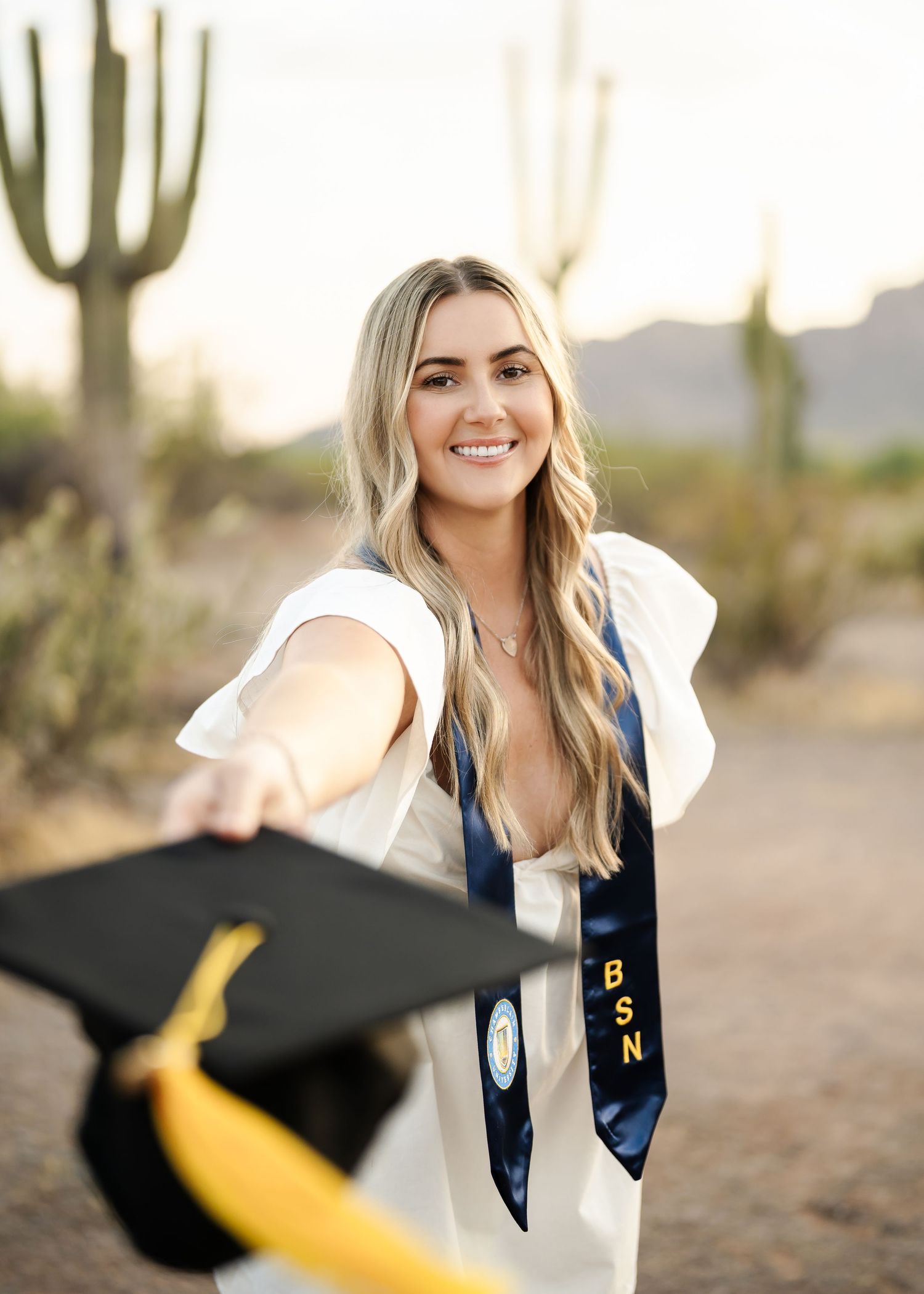 Graduate wearing navy blue stole and tossing cap celebrates in desert landscape with saguaro cacti.