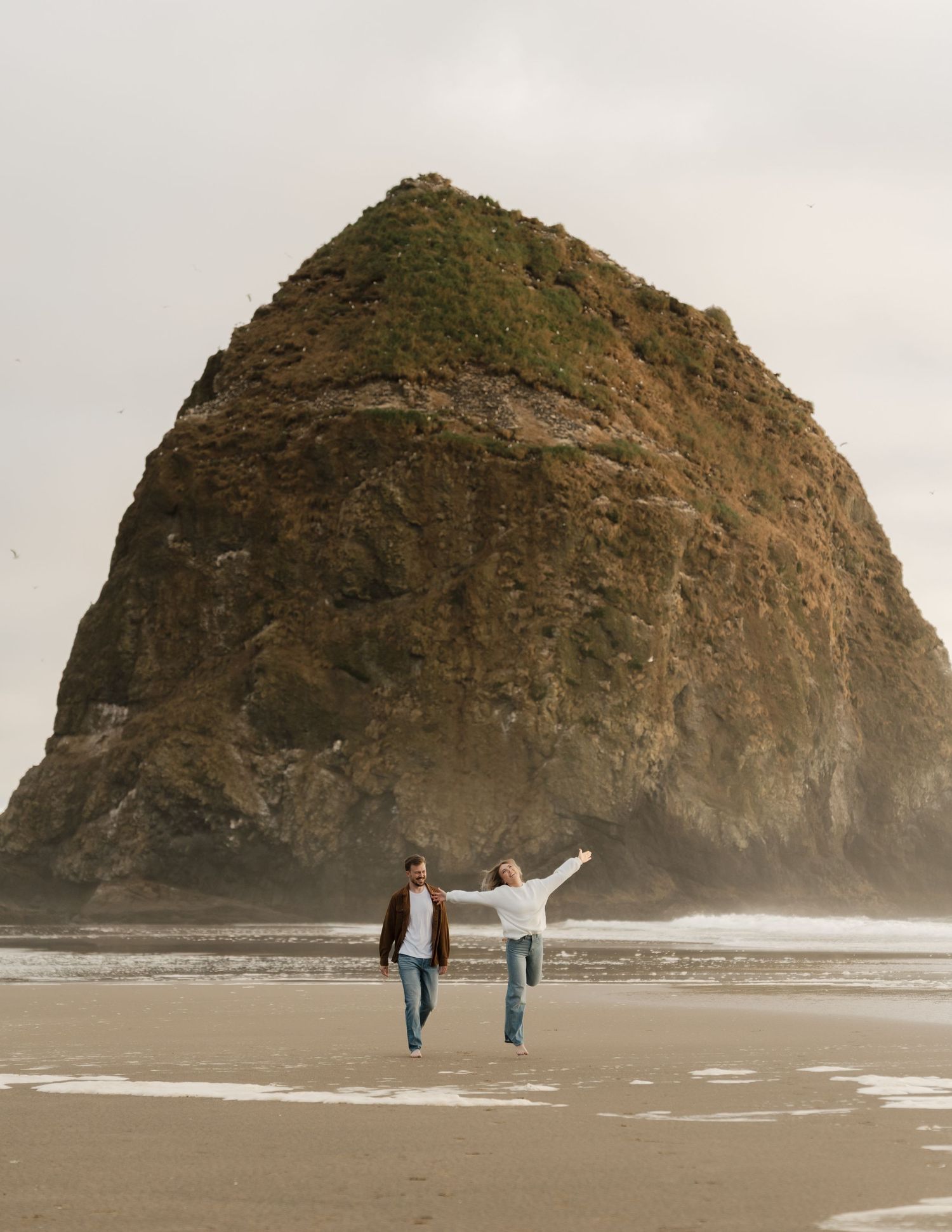 Couple walking hand in hand on Cannon Beach with iconic Haystack Rock in the background on an overcast day.