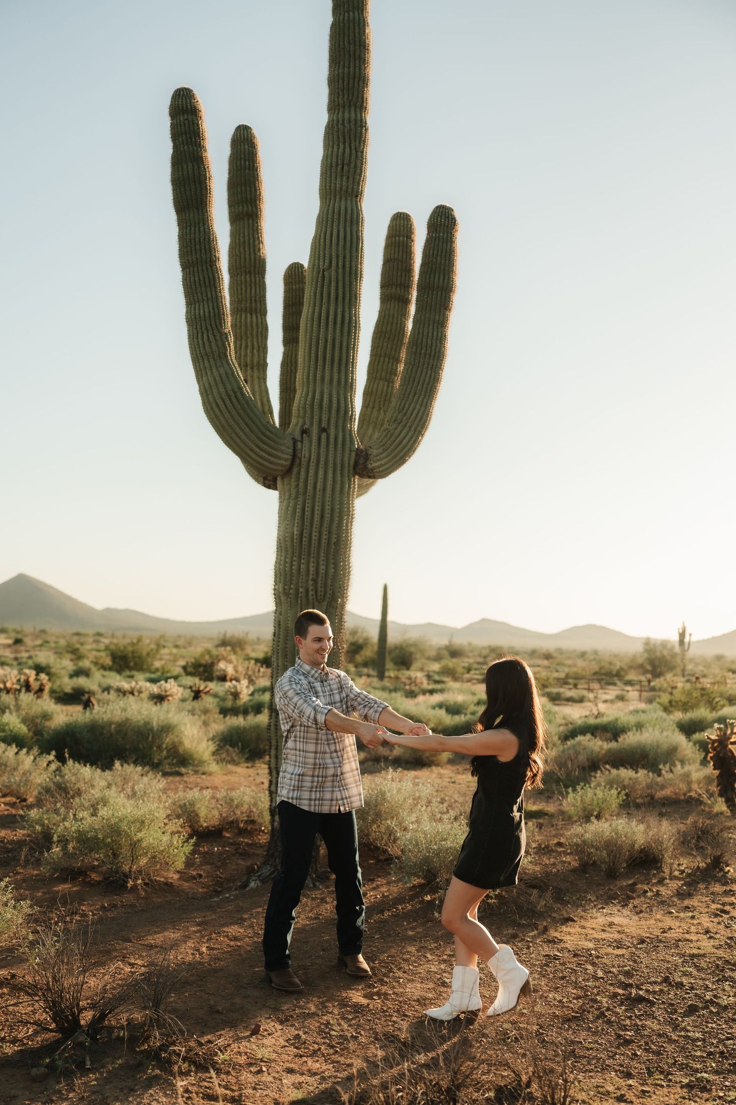 Couple holding hands and dancing in front of a tall saguaro cactus in the Arizona desert at golden hour.