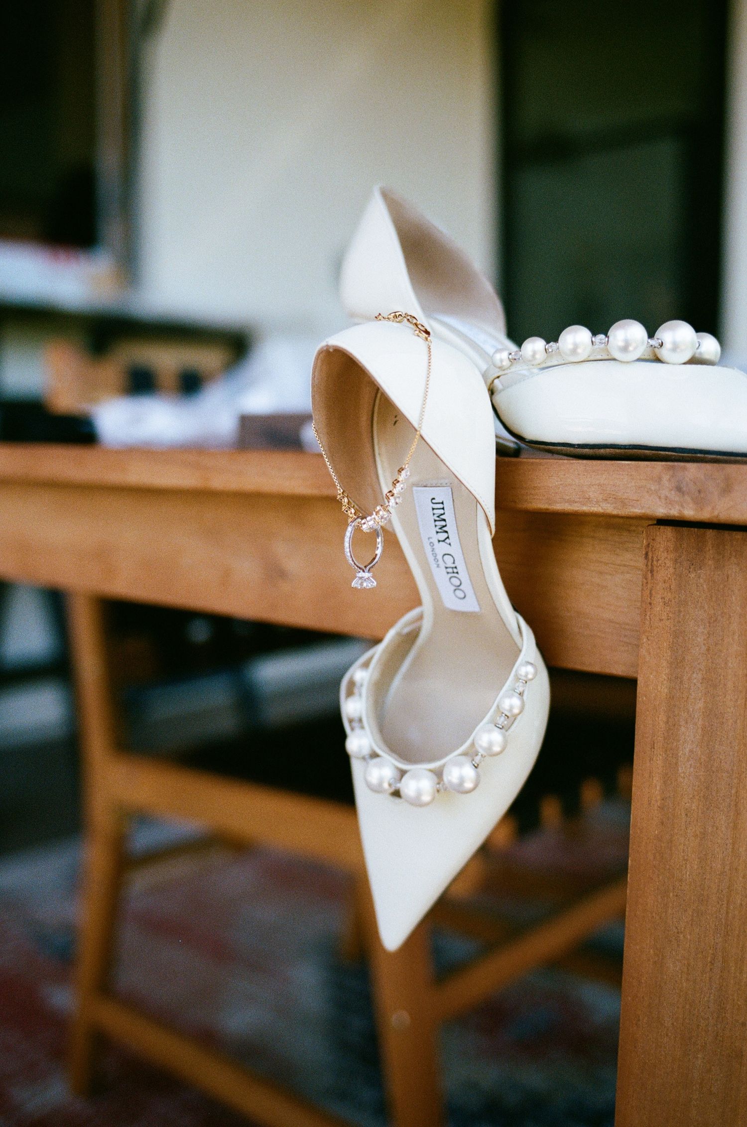 Elegant Jimmy Choo pearl-embellished white bridal heels displayed on a wooden surface with a delicate gold necklace.