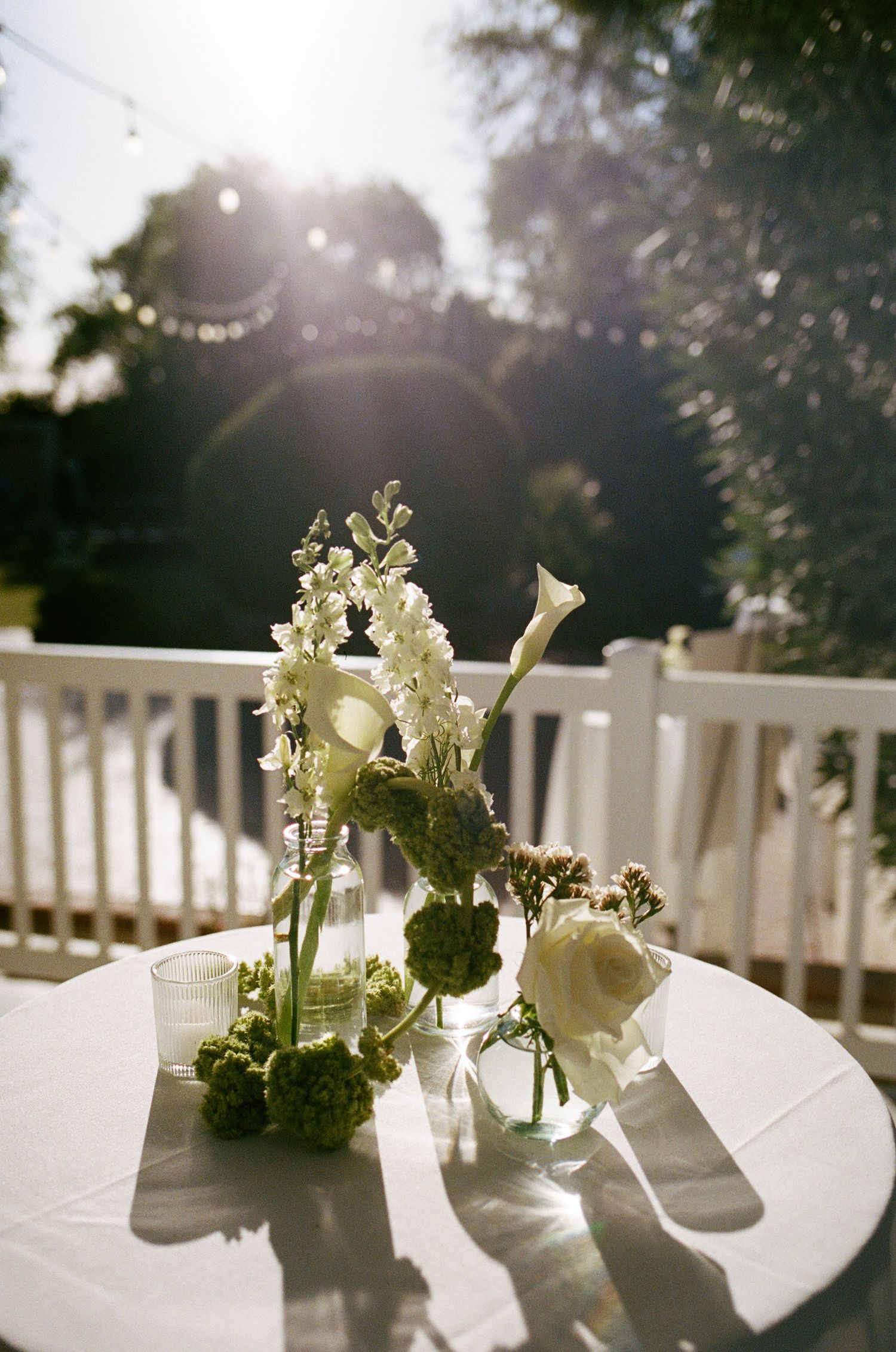 White floral centerpieces in glass vases on a white tablecloth at an outdoor garden wedding reception.