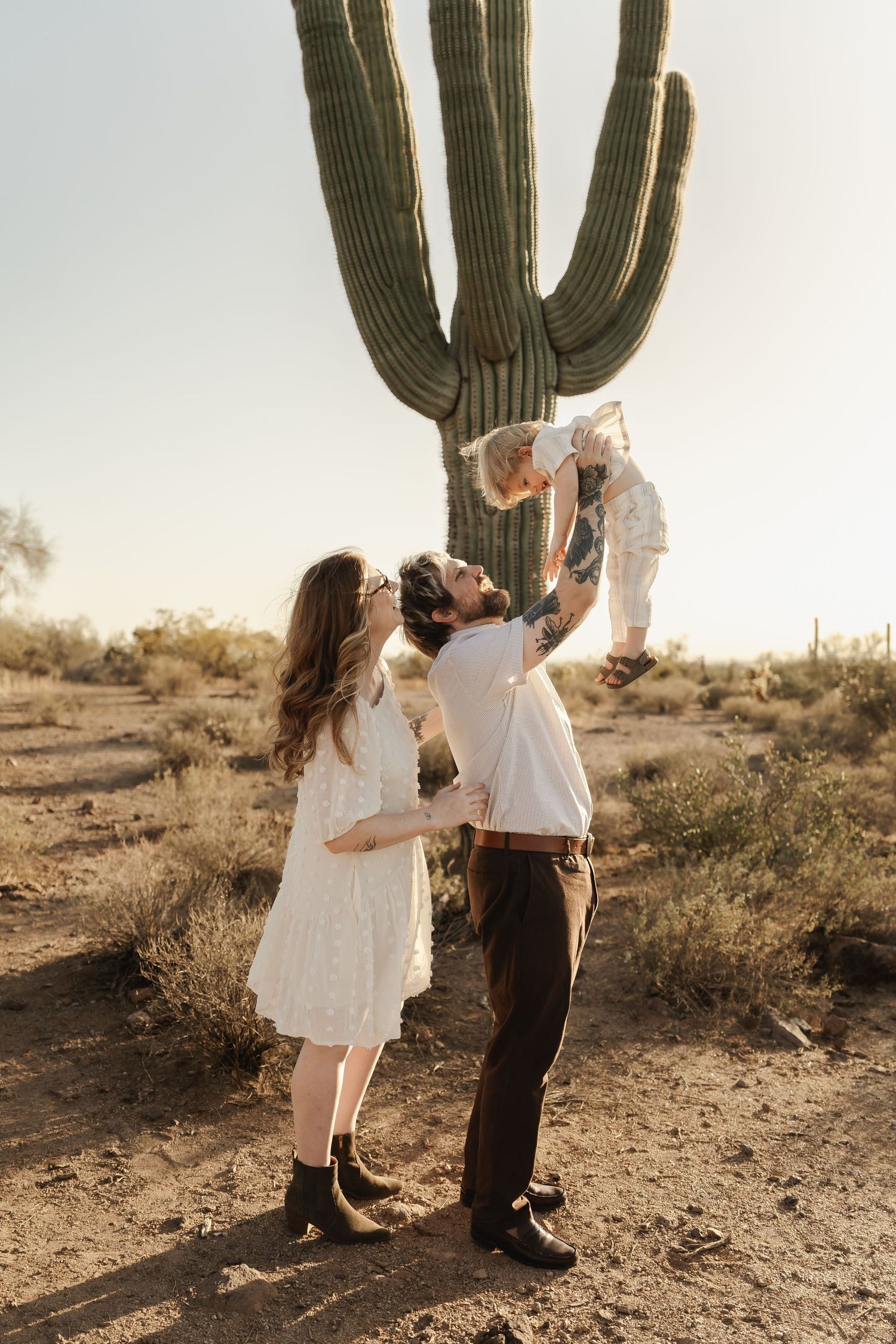 Family members in light colored clothing posing playfully by saguaro cactus at sunset.