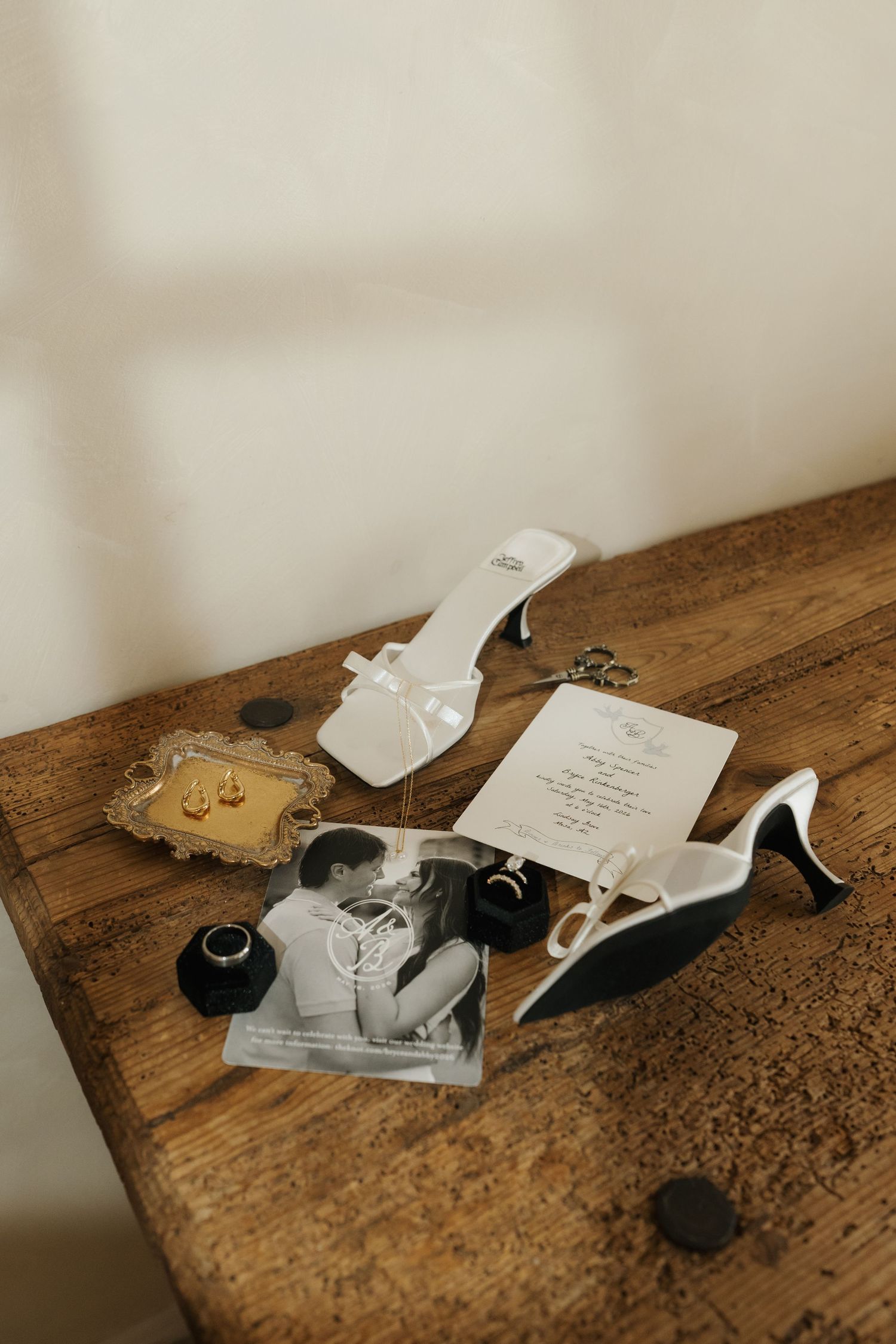 Elegant wedding flat lay with white heels, gold jewelry, rings, invitation card, and black-and-white couple photo on rustic wood table.