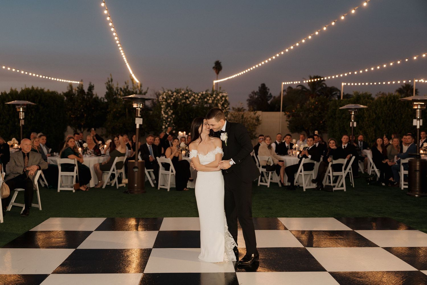 A couple shares their first dance on a checkered dance floor under string lights at an outdoor reception.