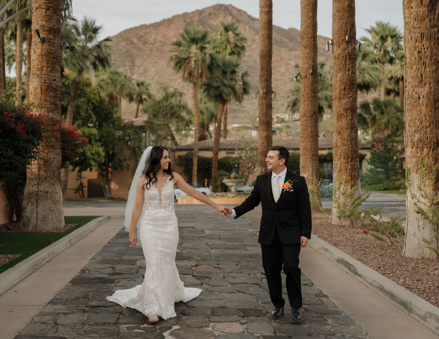 Bride and groom hold hands on a palm-lined path with mountain backdrop at a desert wedding venue.