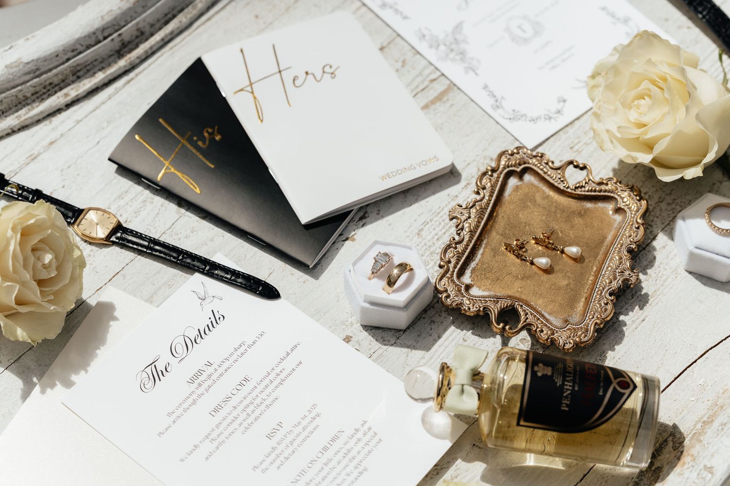 Flat lay of wedding planning items with creamy roses, invitation cards, perfume bottle and ornate gold frame on white surface.
