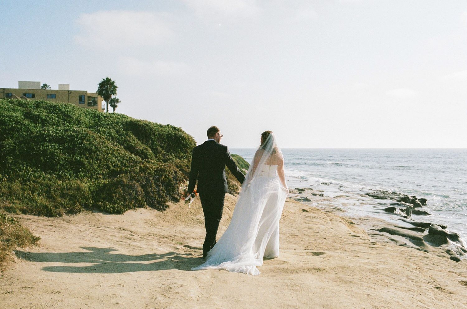 Bride and groom walk hand-in-hand along a sandy coastal bluff overlooking the ocean on their wedding day.
