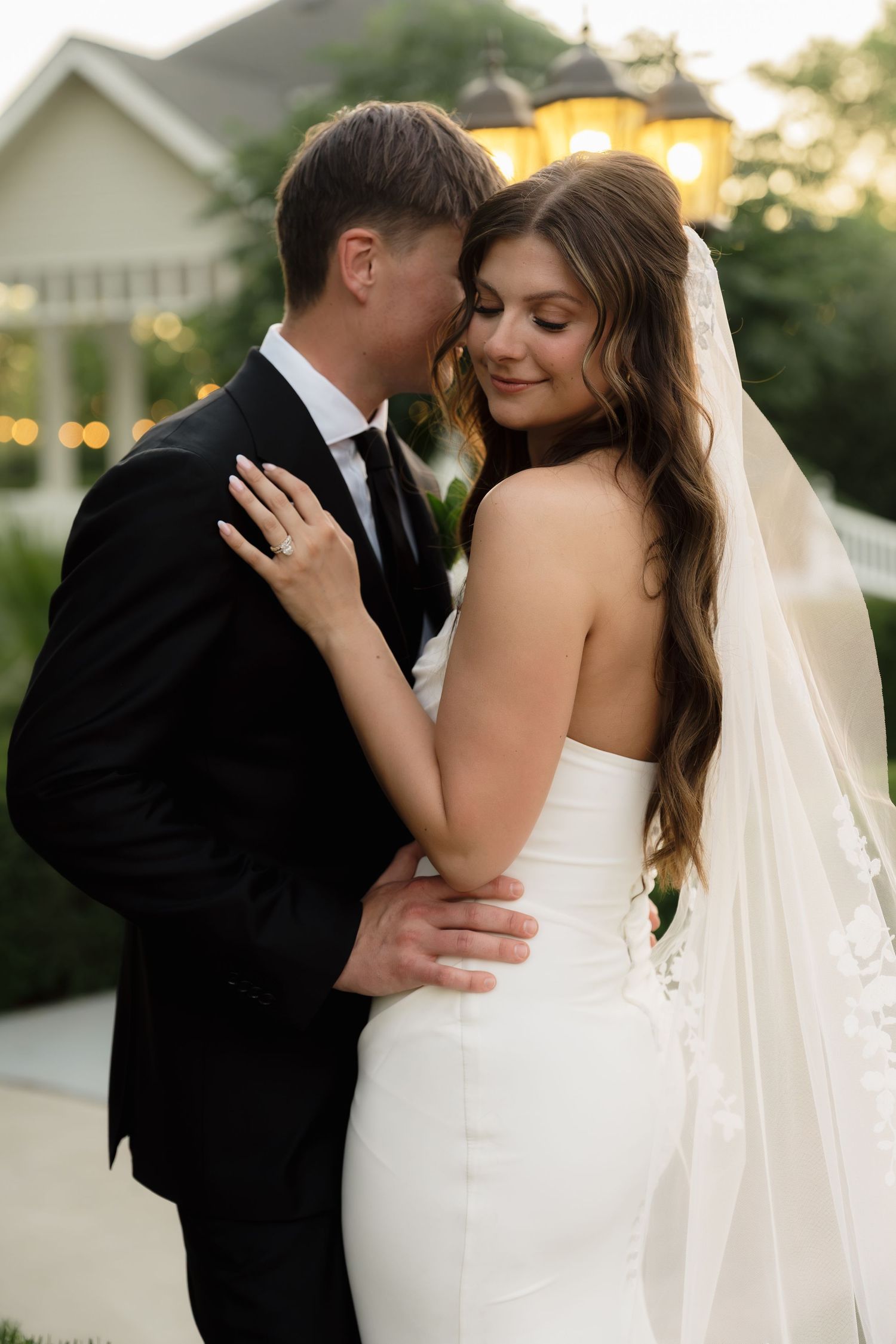 A bride in a white gown and veil embraces her groom in a black suit outdoors at dusk near a warmly lit venue.