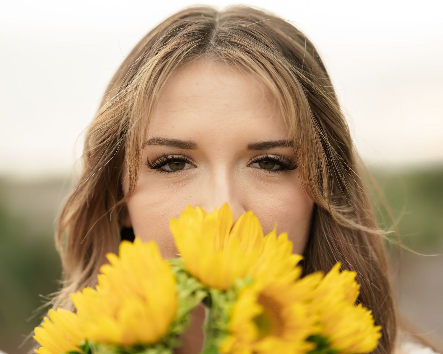 A bright sunflower bouquet positioned in front of someone in a white lace top creates a playful outdoor portrait.