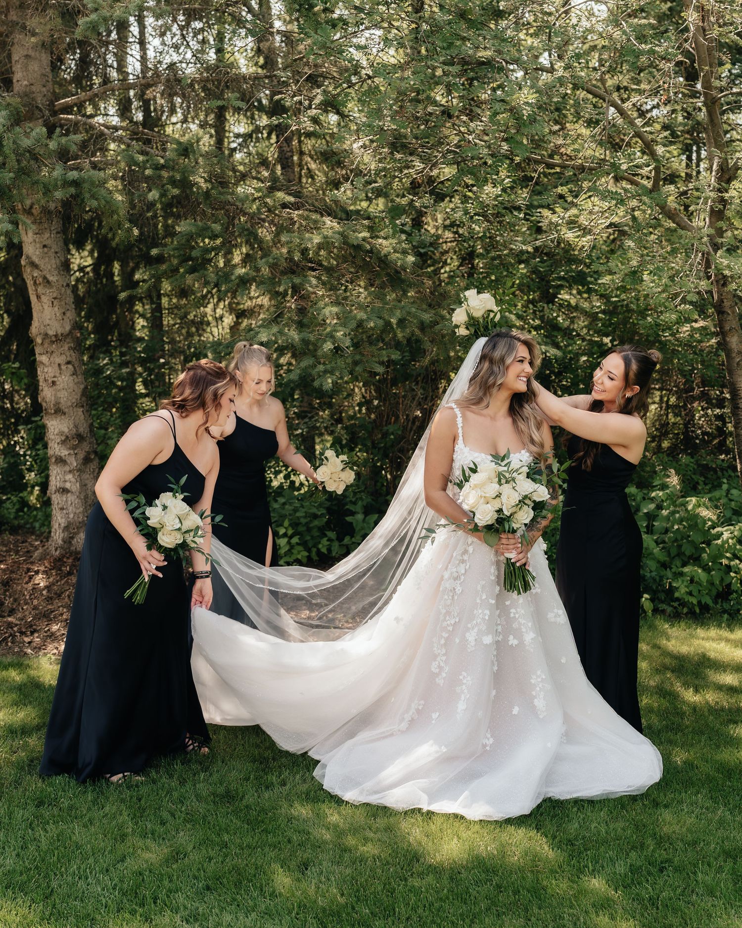 Wedding party helping adjust a flowing white dress train on a sunny lawn with trees in background.