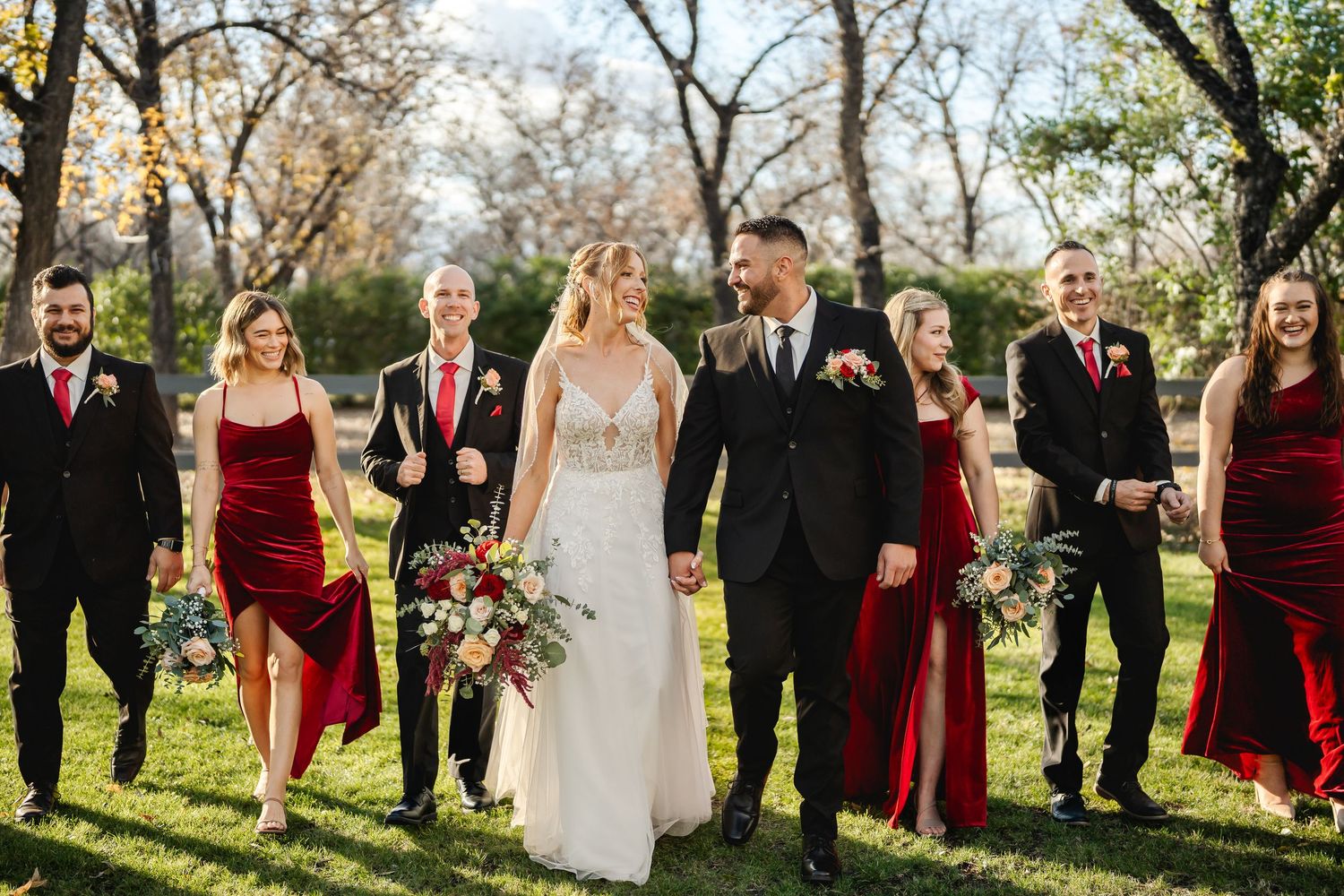 Wedding party in elegant burgundy and black attire pose together outdoors among autumn trees.