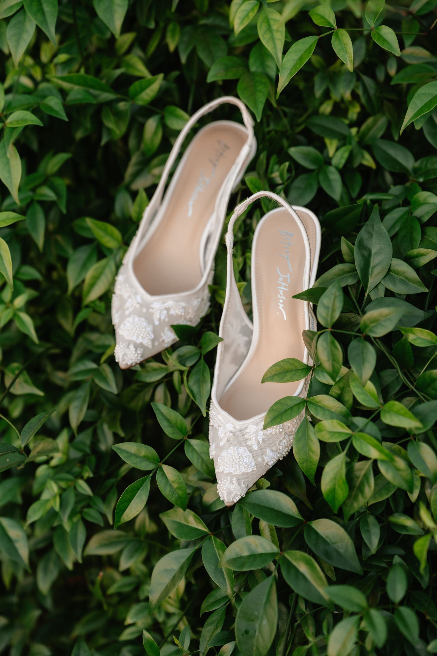 Delicate white lace bridal flats displayed against lush green foliage background.