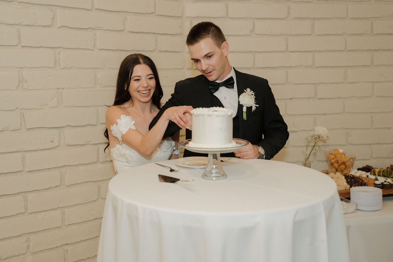 A bride and groom cut their elegant white wedding cake together at a reception with a white brick wall backdrop.