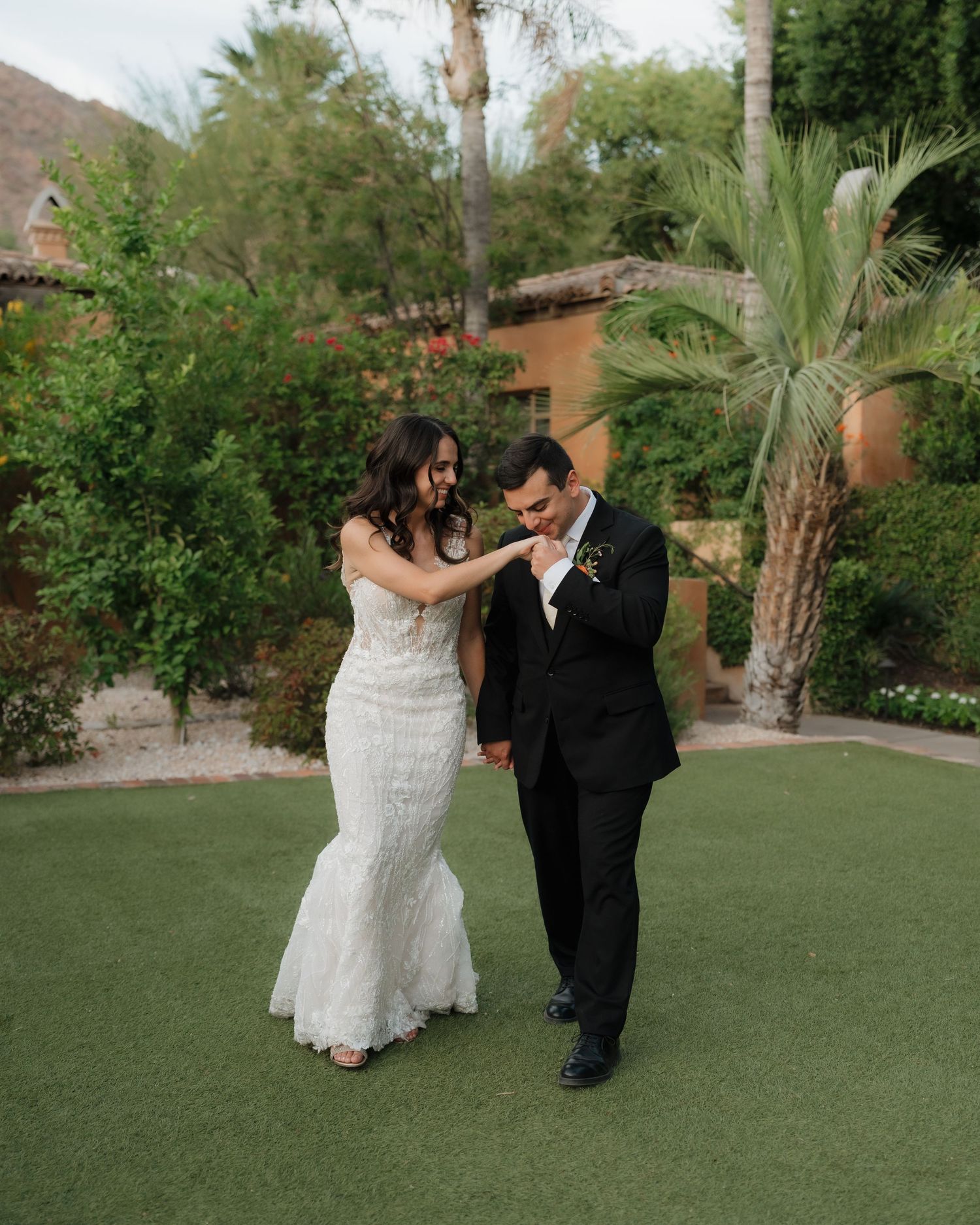 Groom kissing bride's hand in a lush garden setting, both dressed in elegant wedding attire.