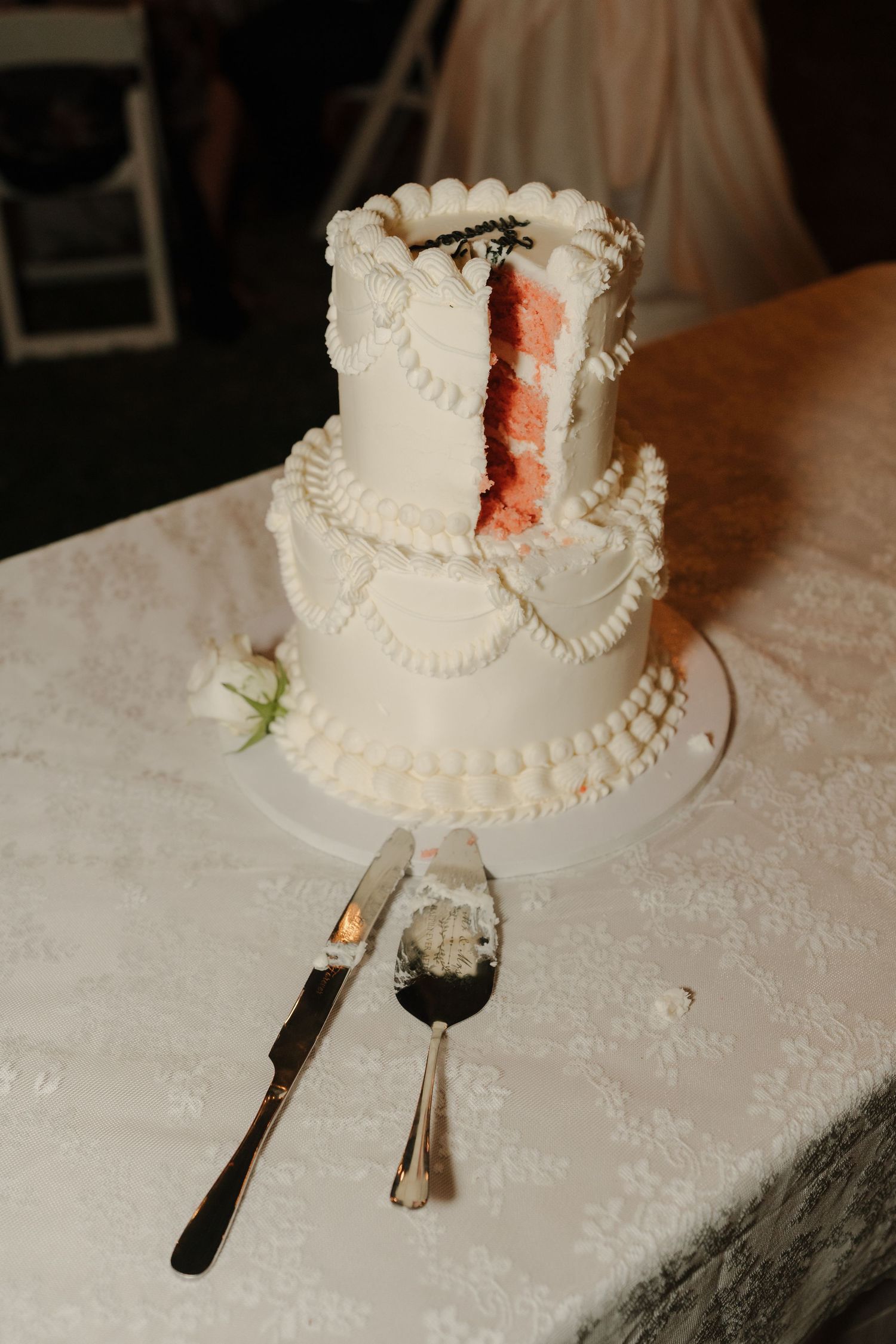 Two-tier white wedding cake with pearl decorations, partially sliced to reveal pink interior, displayed on a lace-covered table.