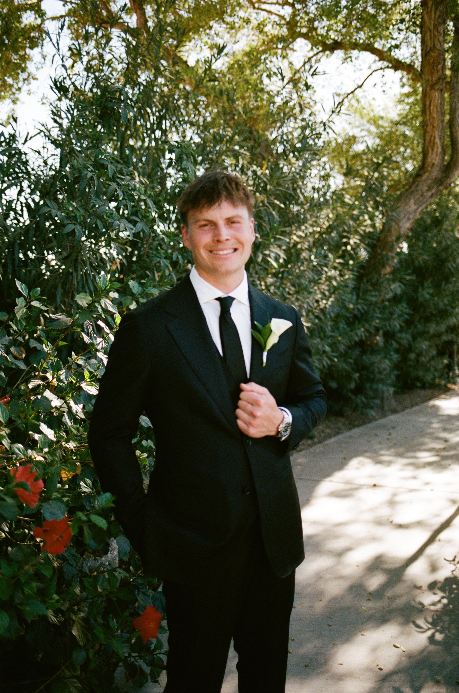 Smiling groom in black suit with white boutonniere standing on garden path surrounded by lush green trees.