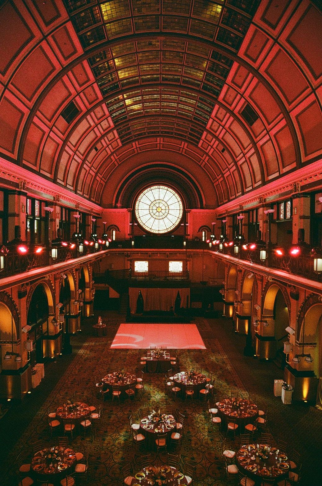 Grand ballroom with arched red ceiling, round stained glass window, elegant round tables, and a glowing dance floor.
