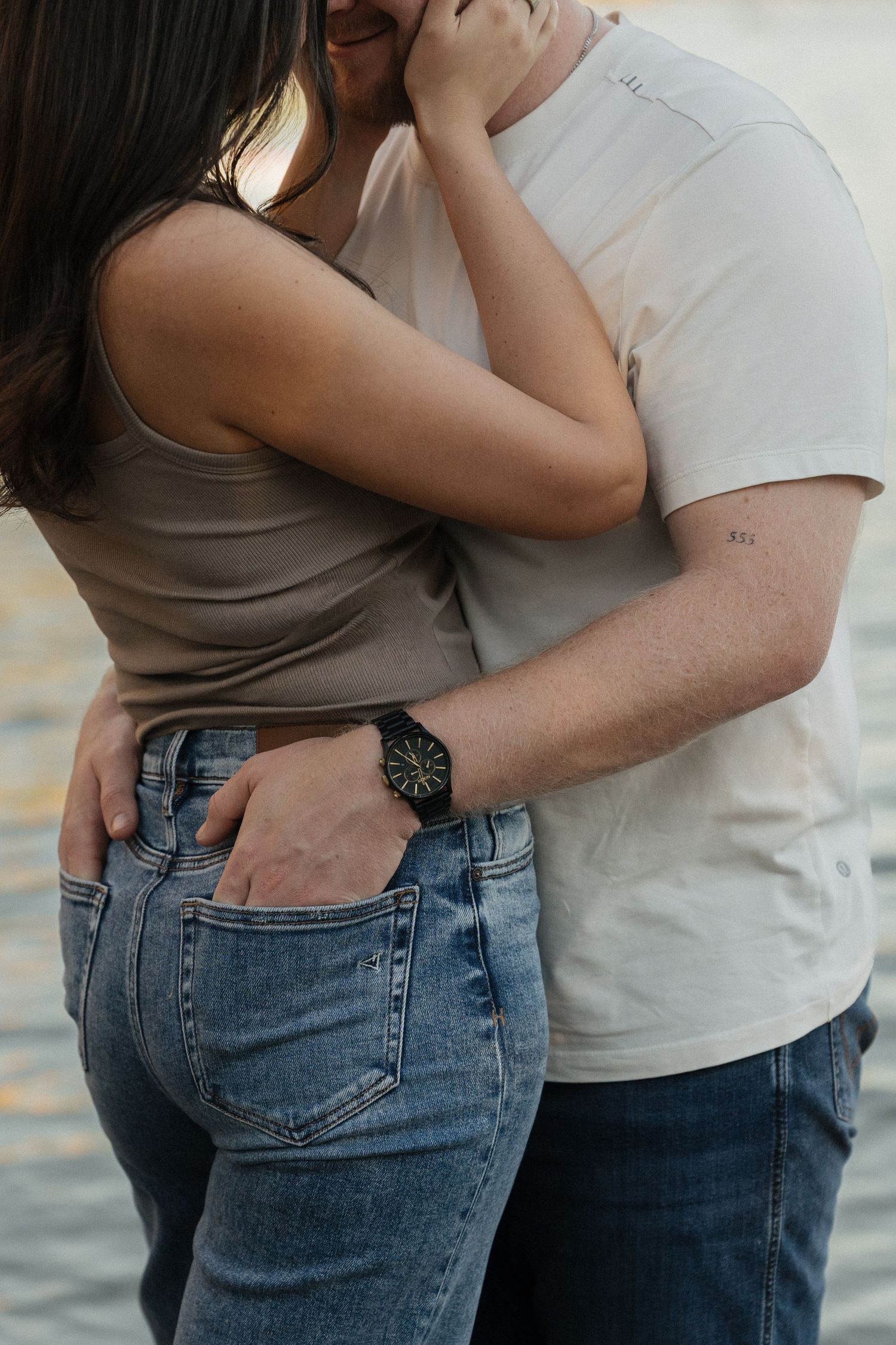 Couple embracing intimately, man wearing black watch and white t-shirt, woman in beige tank top and blue jeans.