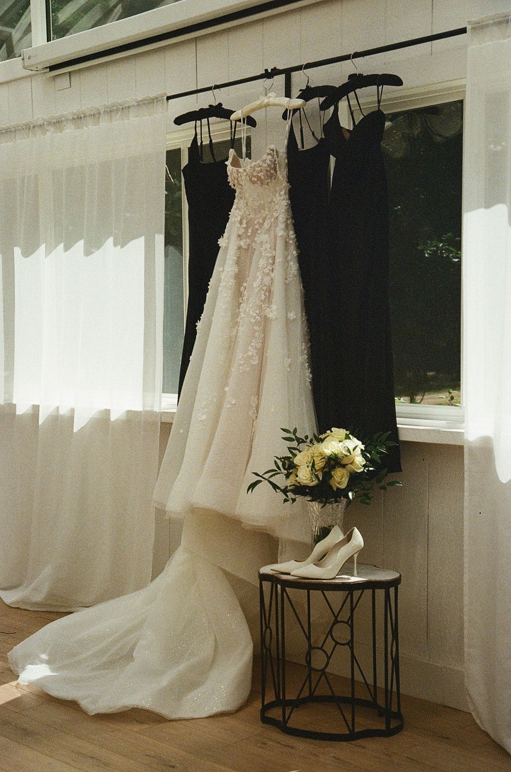 Elegant wedding dress with floral details hanging beside dark bridesmaid gowns, with white heels and bouquet on a side table.