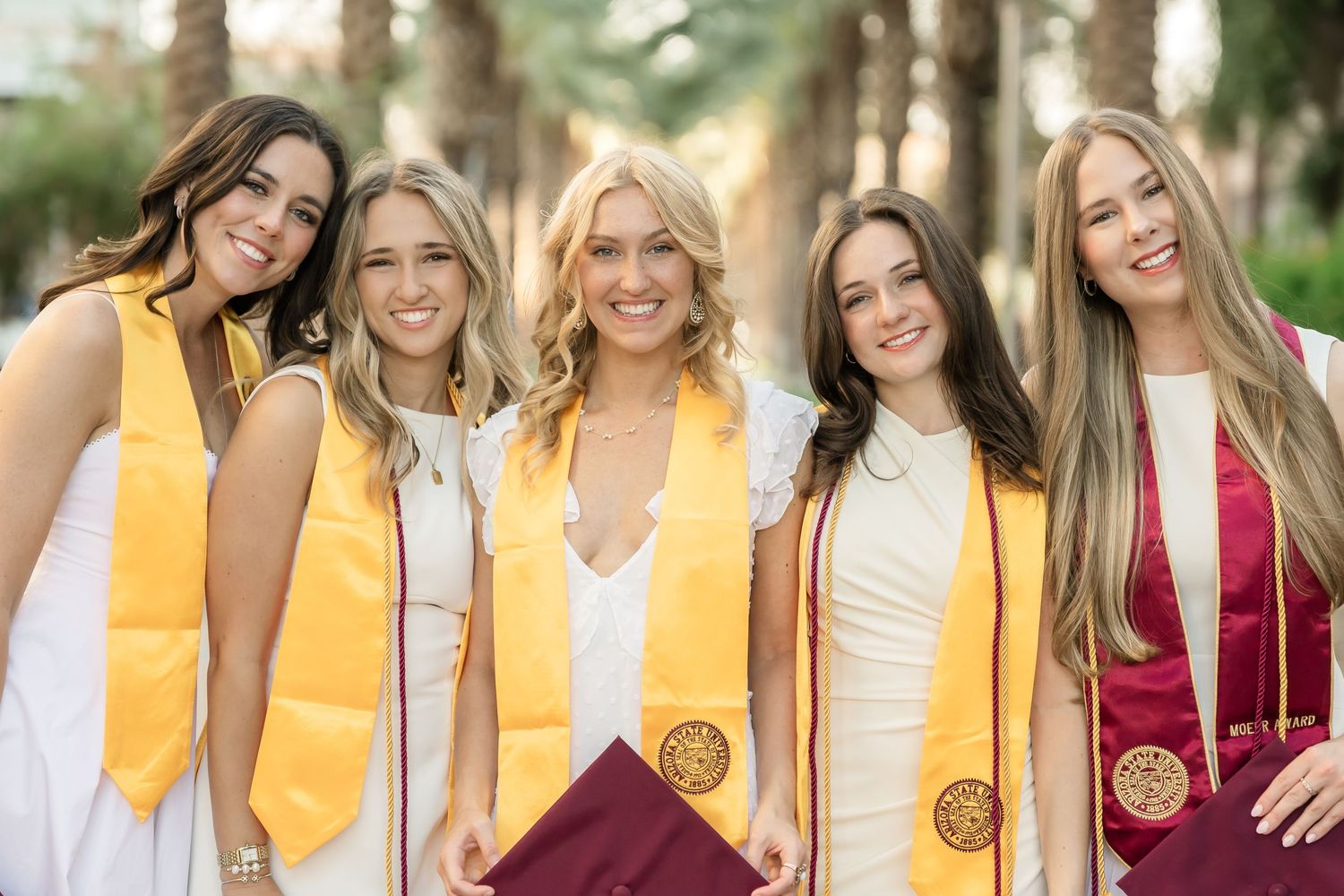 Graduates in maroon gowns and gold stoles pose together in friendship circle.