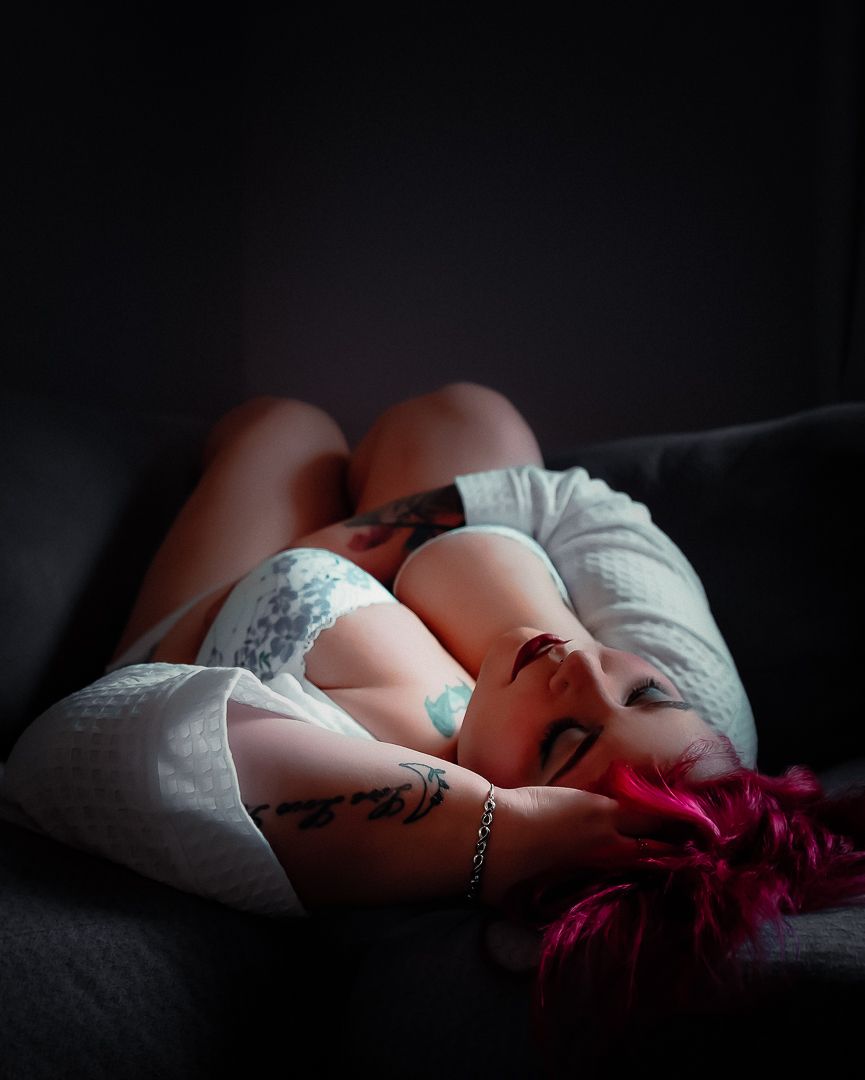 Woman with pink hair and tattoos resting on a dark sofa, wearing a white top and lace lingerie.