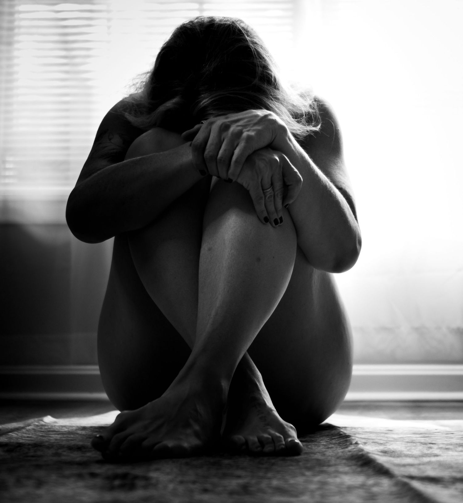 Black and white photo of a woman sitting on floor hugging knees in a vulnerable, emotional pose near a window.