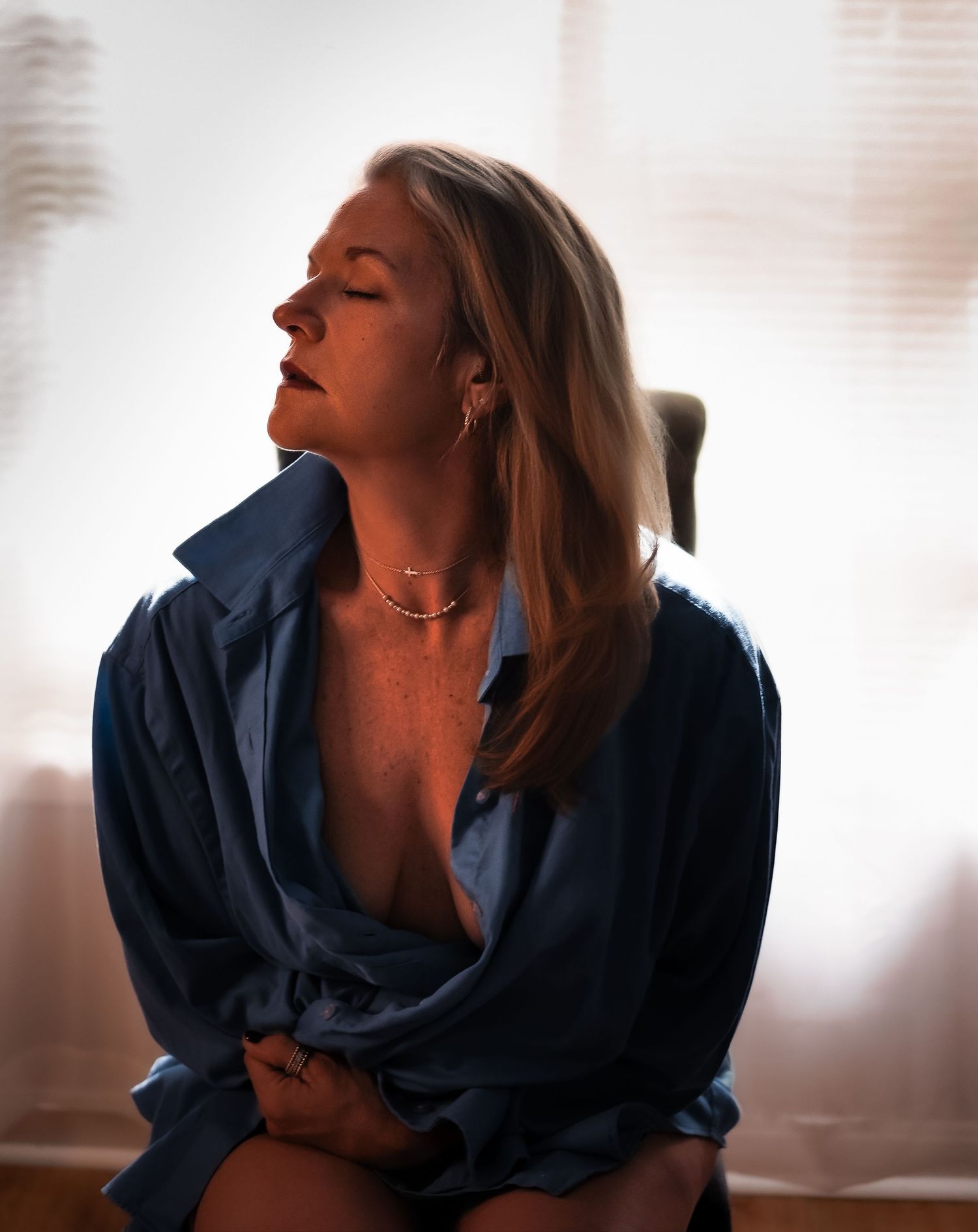 Woman in open blue shirt seated by window with soft backlight, eyes closed, wearing gold necklace and earrings.