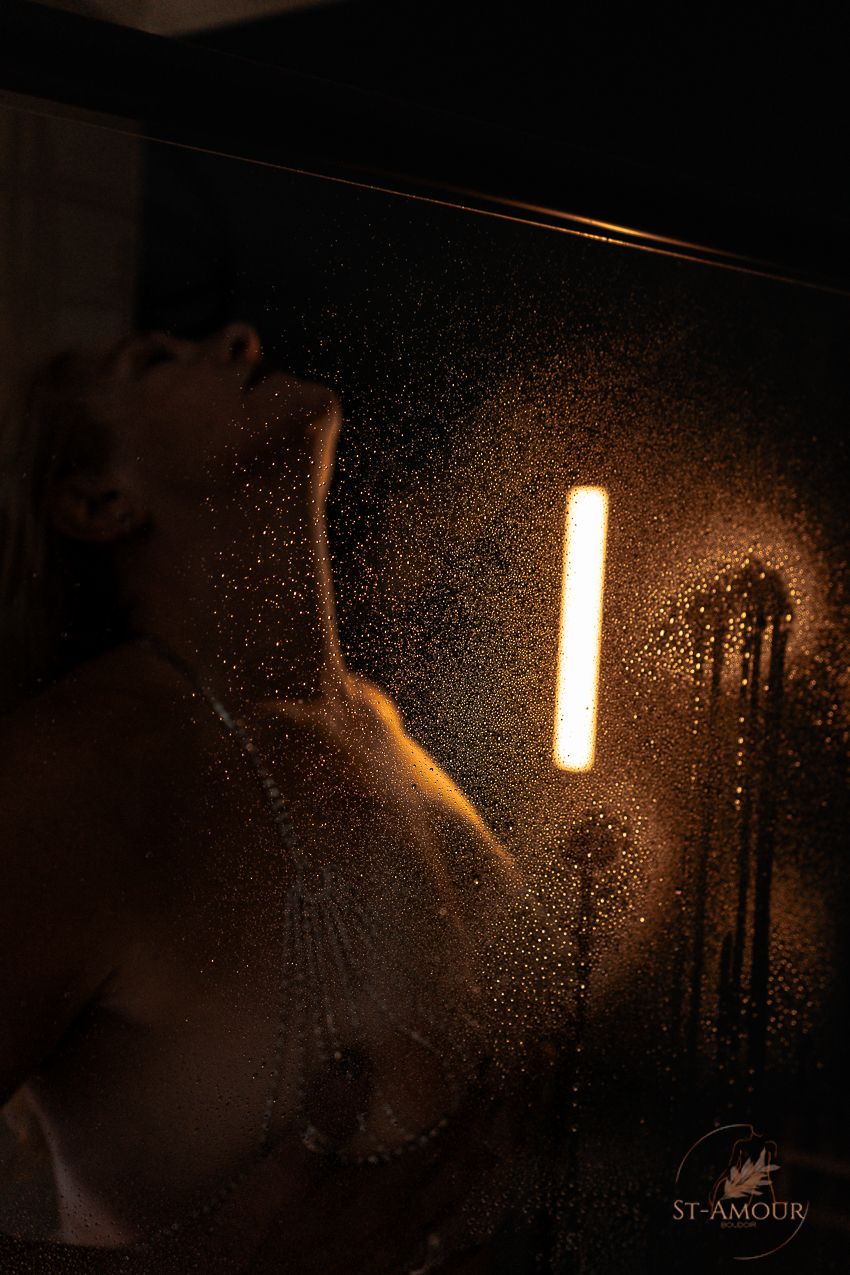 A silhouette of a woman behind a wet glass shower door illuminated by warm golden light.