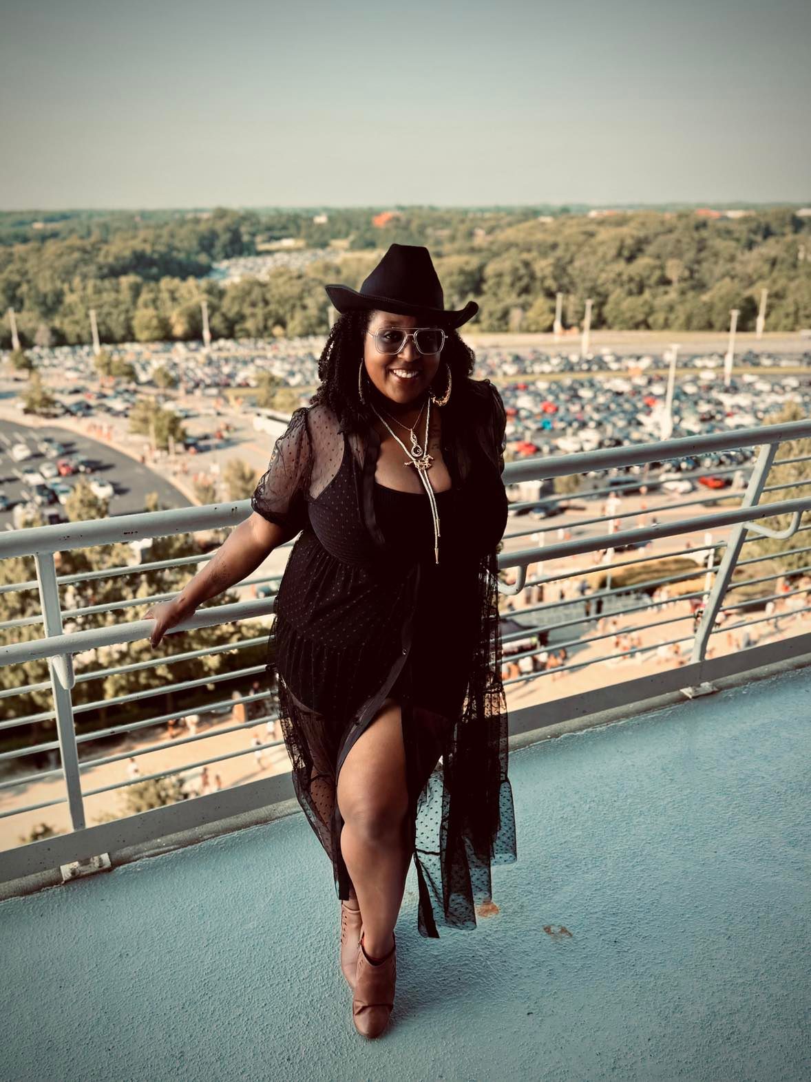 A person in a dark outfit photographed sitting on an elevated balcony with a city view below against an urban backdrop.