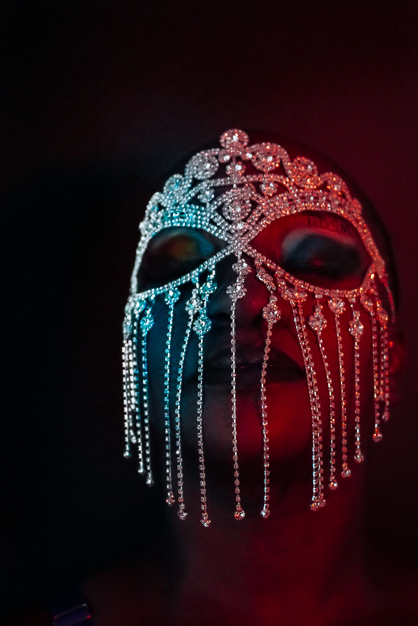 Ornate crystal mask with hanging jewels illuminated by dramatic red and blue lighting against dark background.