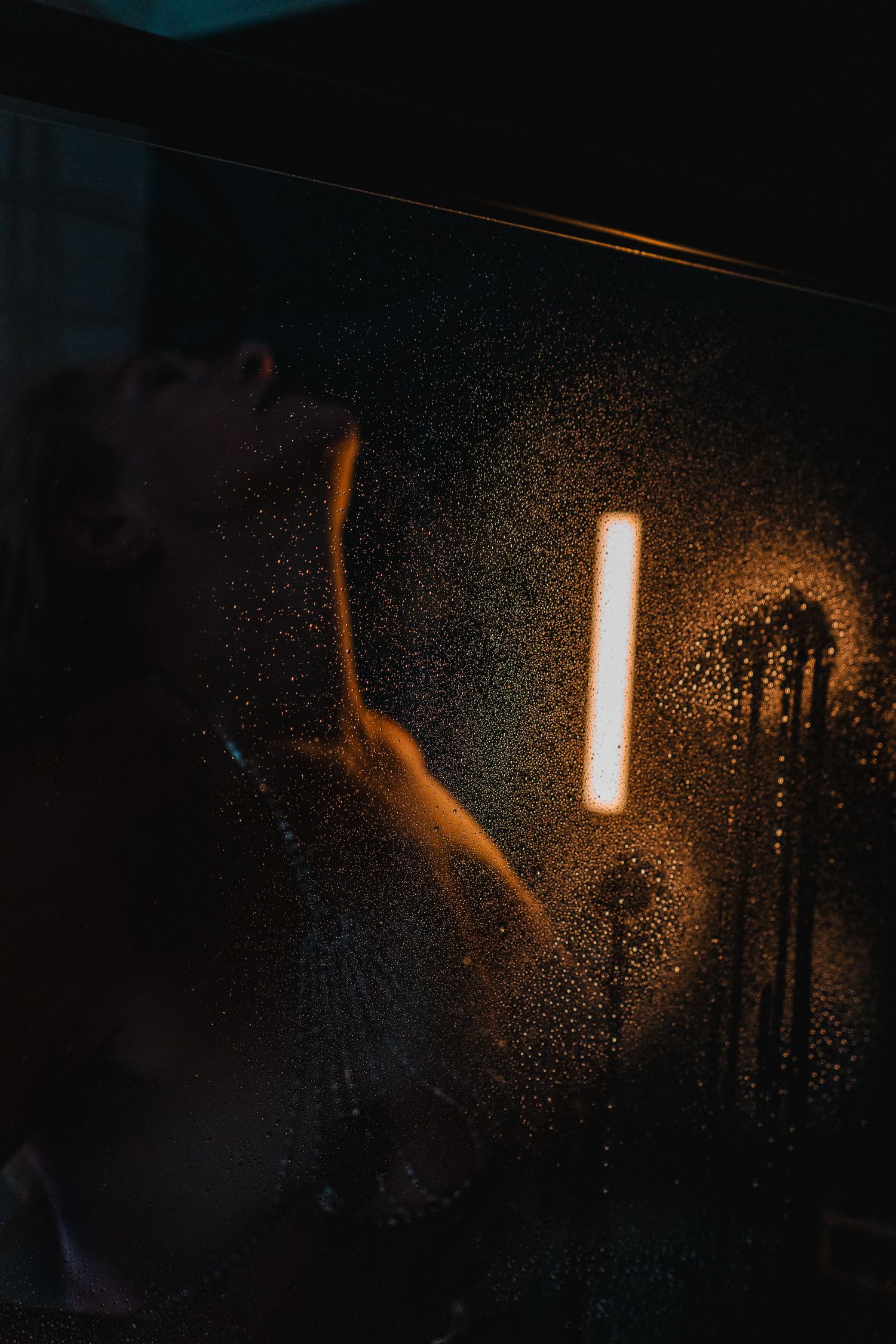 A glowing light illuminates water droplets on a dark window surface creating a moody atmospheric effect.