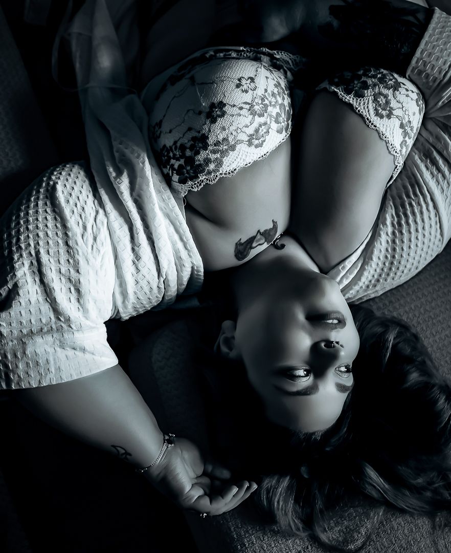 Dramatic black and white boudoir photo showing figure in lace lingerie laying on fabric with high contrast lighting.