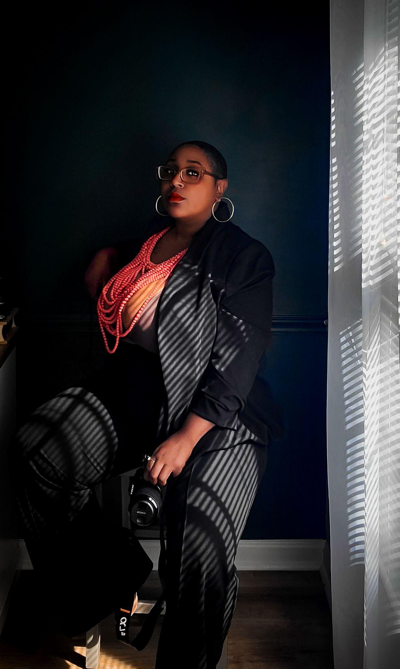 Dramatic portrait with venetian blinds casting shadow patterns across a figure in dark room wearing coral necklaces.
