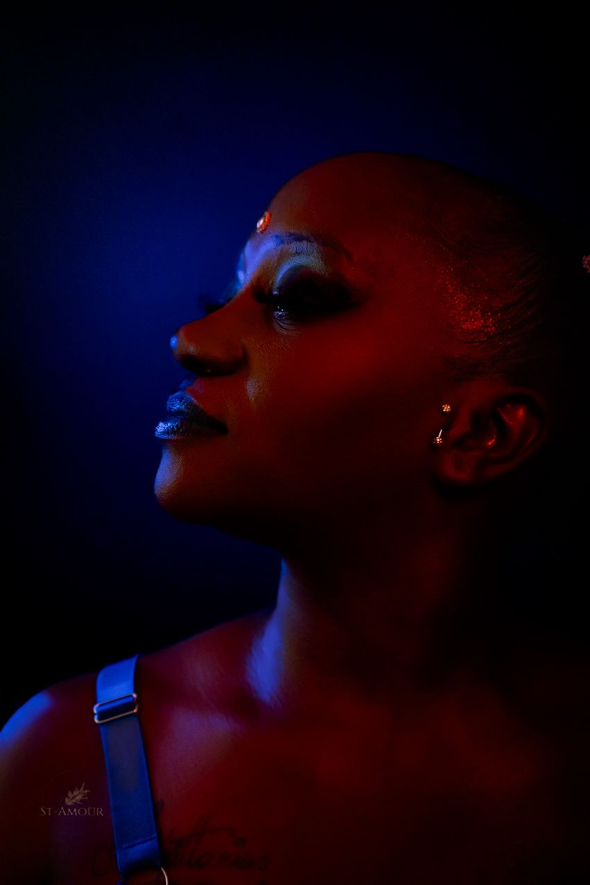 A bald person with dramatic makeup poses in profile against a dark background with blue and red lighting.