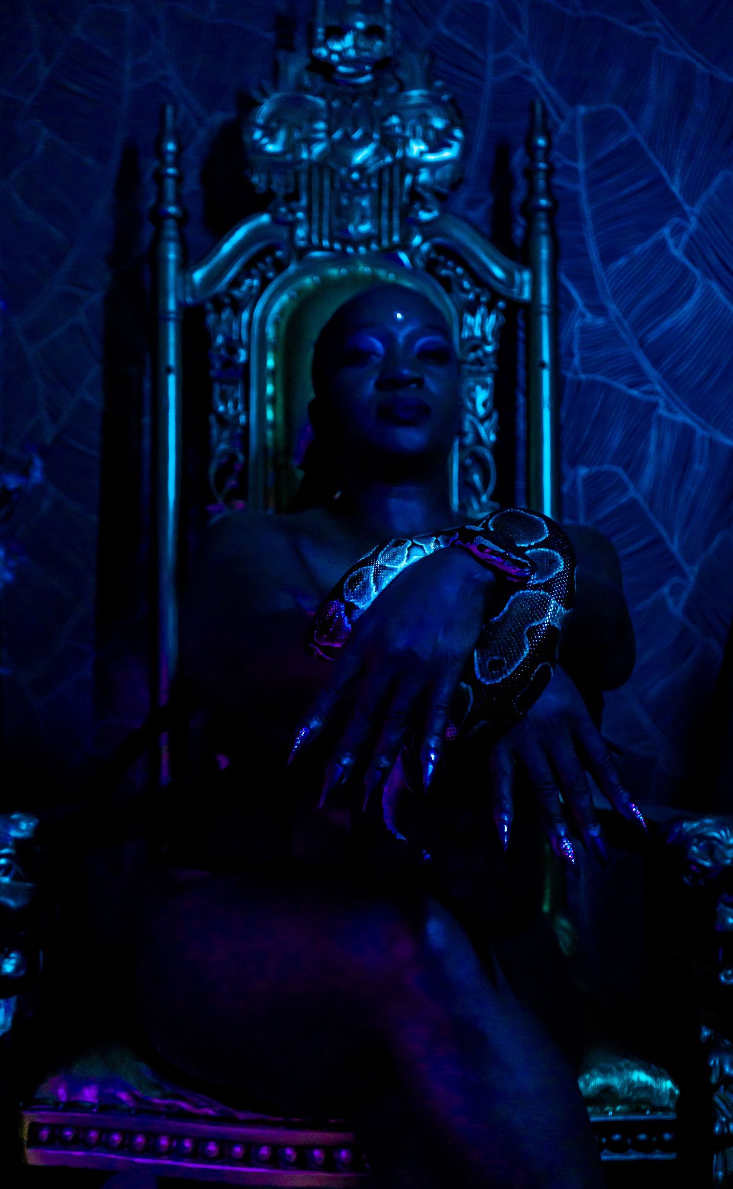 A person sits on an ornate throne bathed in moody blue lighting while holding what appears to be a glowing object.