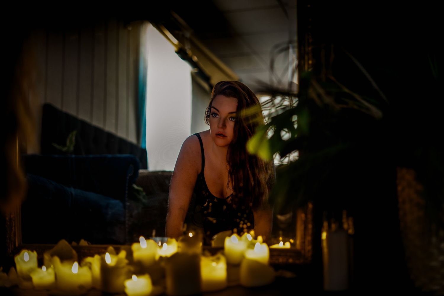 Woman in black dress surrounded by glowing candles in a dimly lit room with moody, atmospheric lighting.