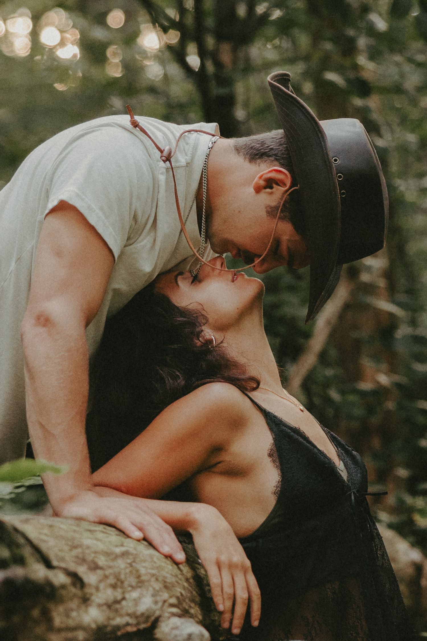 A couple shares a romantic kiss in a forest, man wearing a cowboy hat leans down to kiss a woman in a black dress.