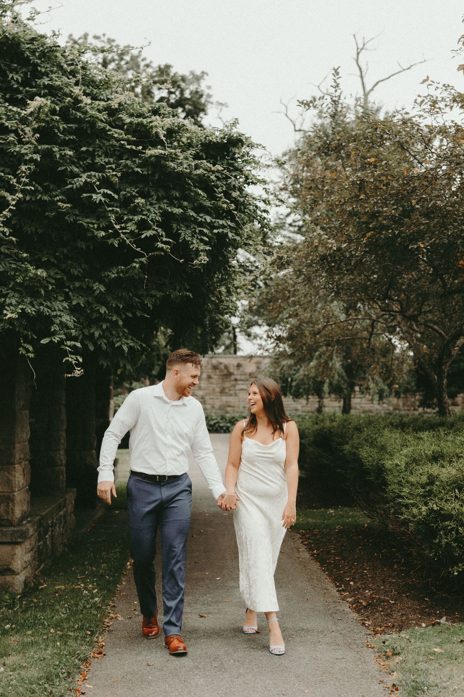 Couple holding hands walking along a garden path, woman in white dress and man in blue pants and white shirt.