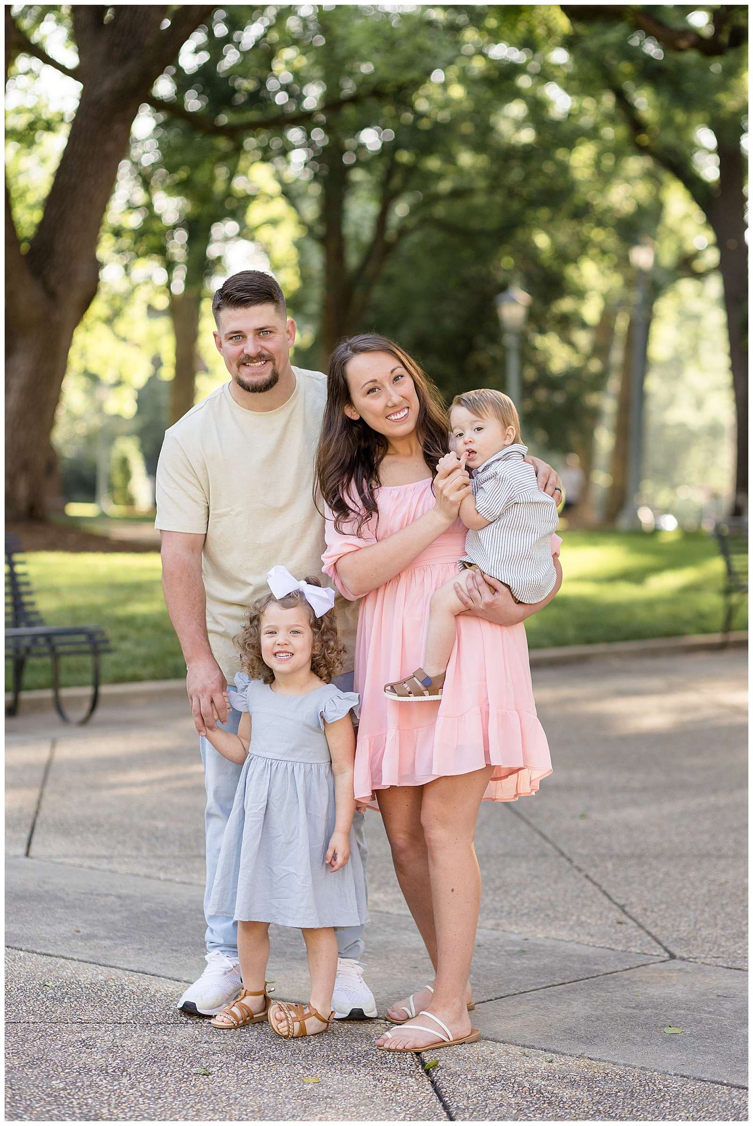 Family of four poses outdoors in a park, parents with toddler girl and baby boy, wearing pastel outfits for Photos in DT Raleigh NC