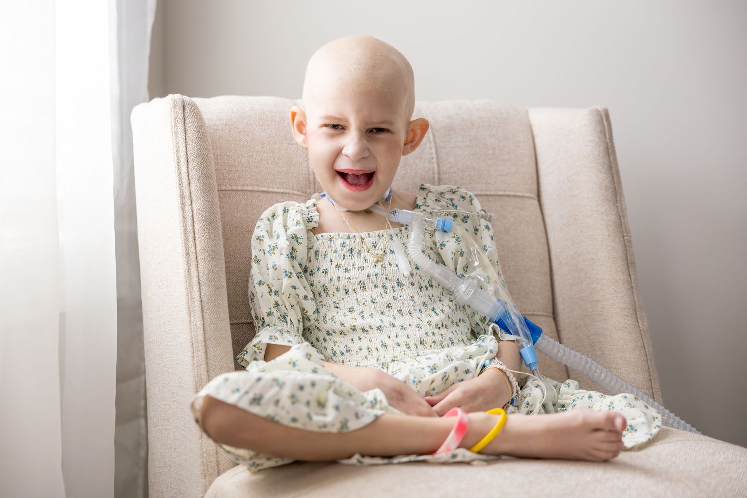 Young girl wearing floral dress sits cross-legged on beige chair while holding her hands together and smiling.